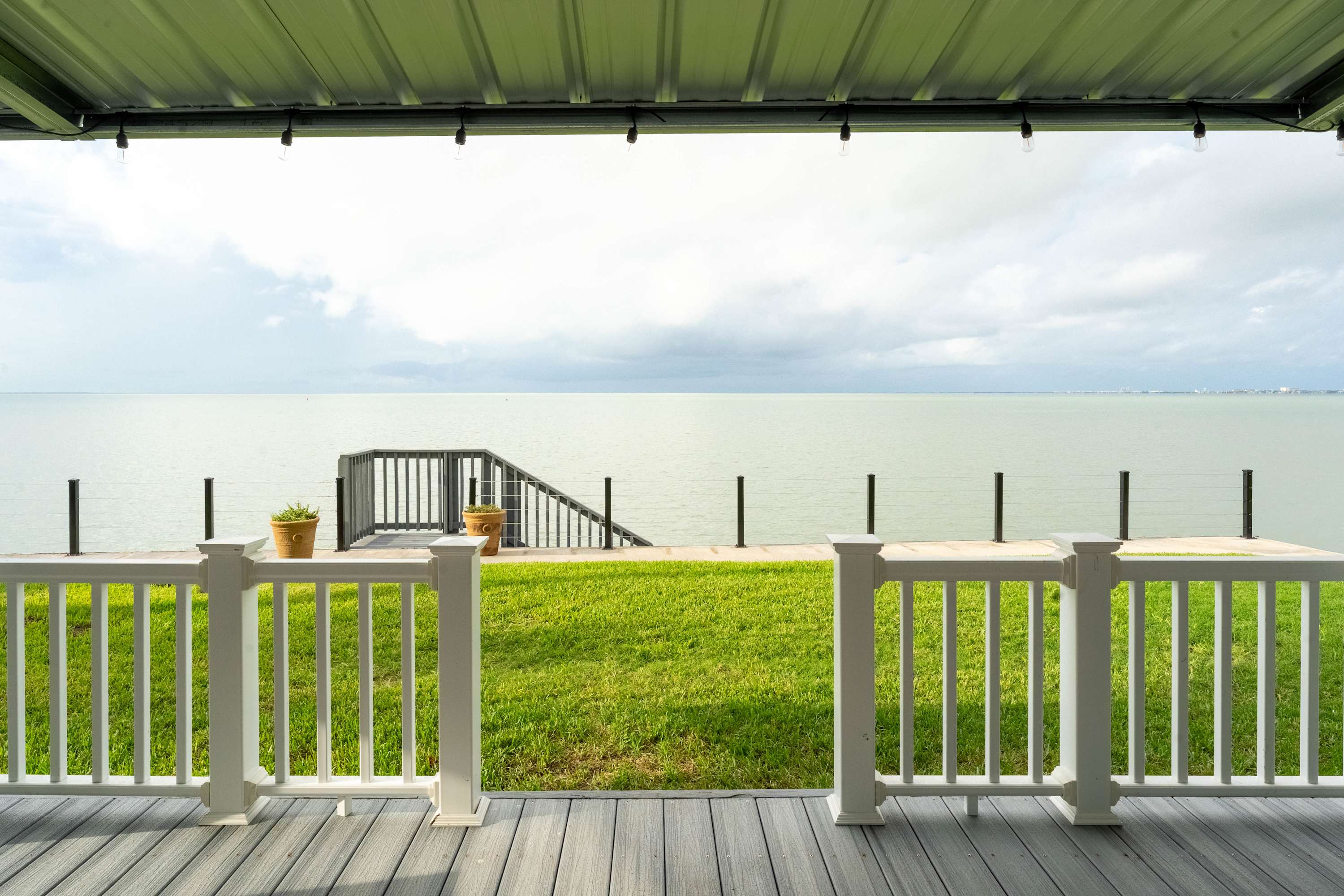 A view from a covered deck looking out over a green lawn to the calm bay water, with a set of stairs leading down to the water. White railings frame the deck, and string lights are visible overhead.
