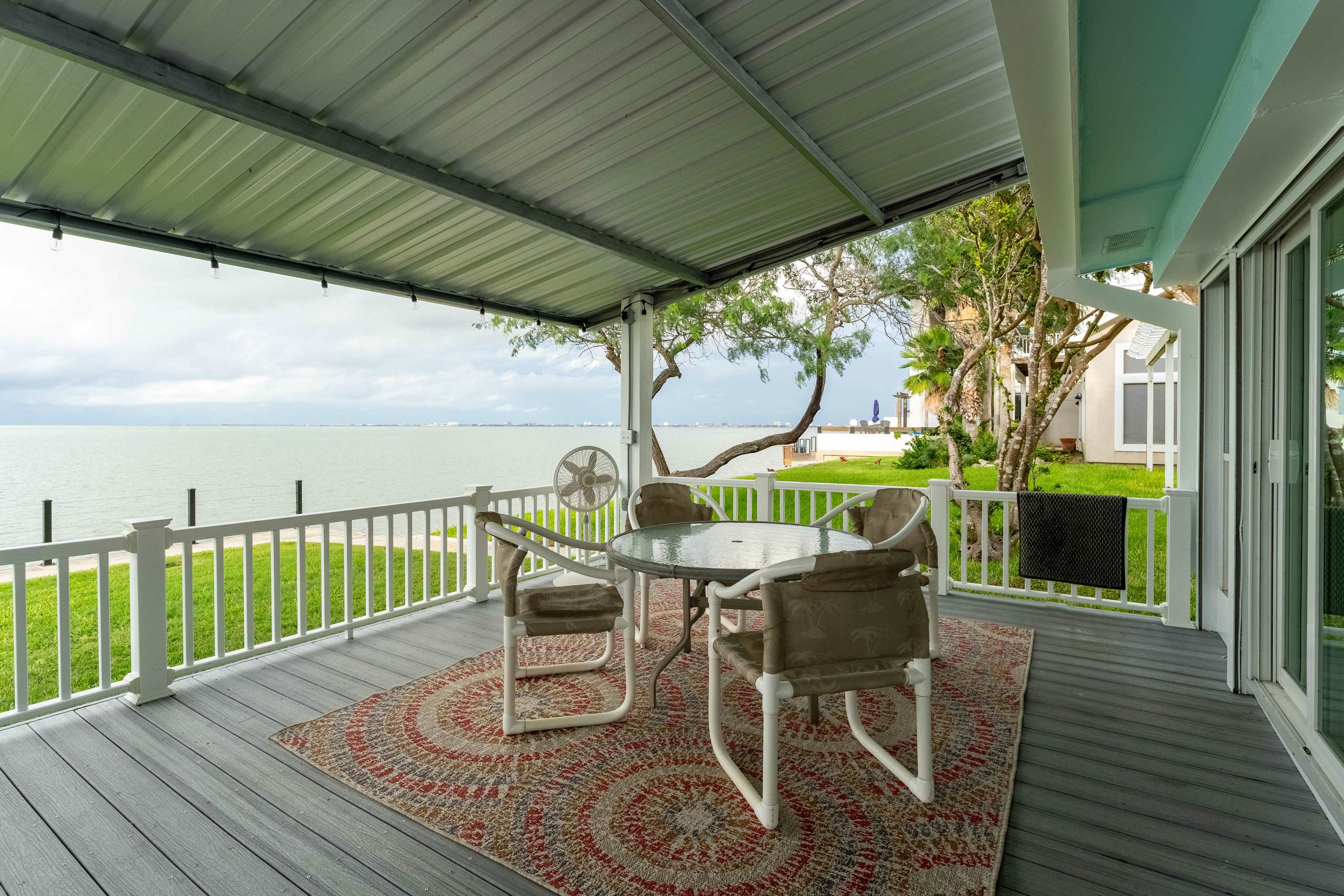 A covered outdoor patio with a gray deck, featuring a round glass-top dining table with four chairs and a colorful circular rug. A white railing separates the patio from a grassy lawn and the calm bay water beyond.