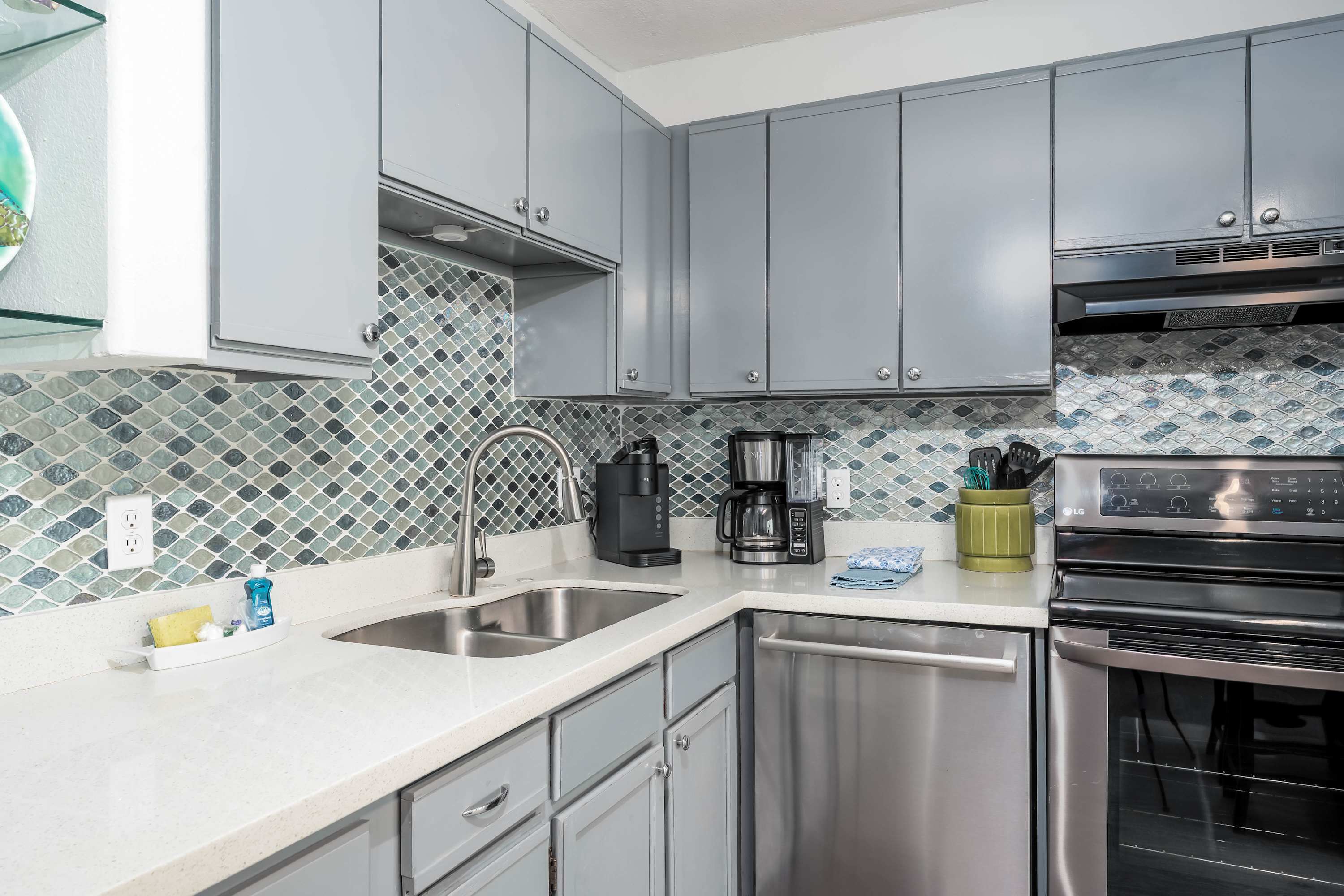 A modern kitchen features light gray cabinets, a white countertop, and a blue and green mosaic tile backsplash, equipped with a stainless steel sink, dishwasher, and black appliances.