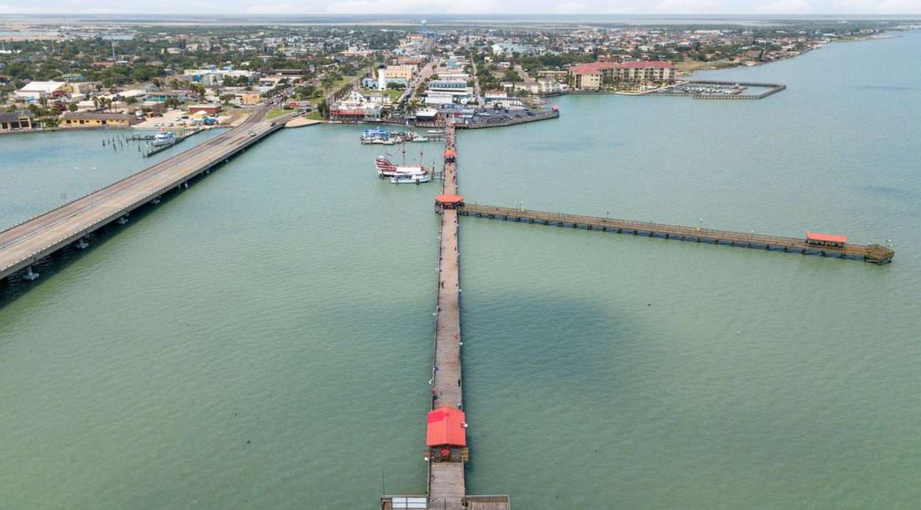 An aerial view captures a very long pier extending far into the calm bay, with a road bridge running parallel to the shoreline. A bustling town with buildings and boats is visible along the waterfront in the distance.