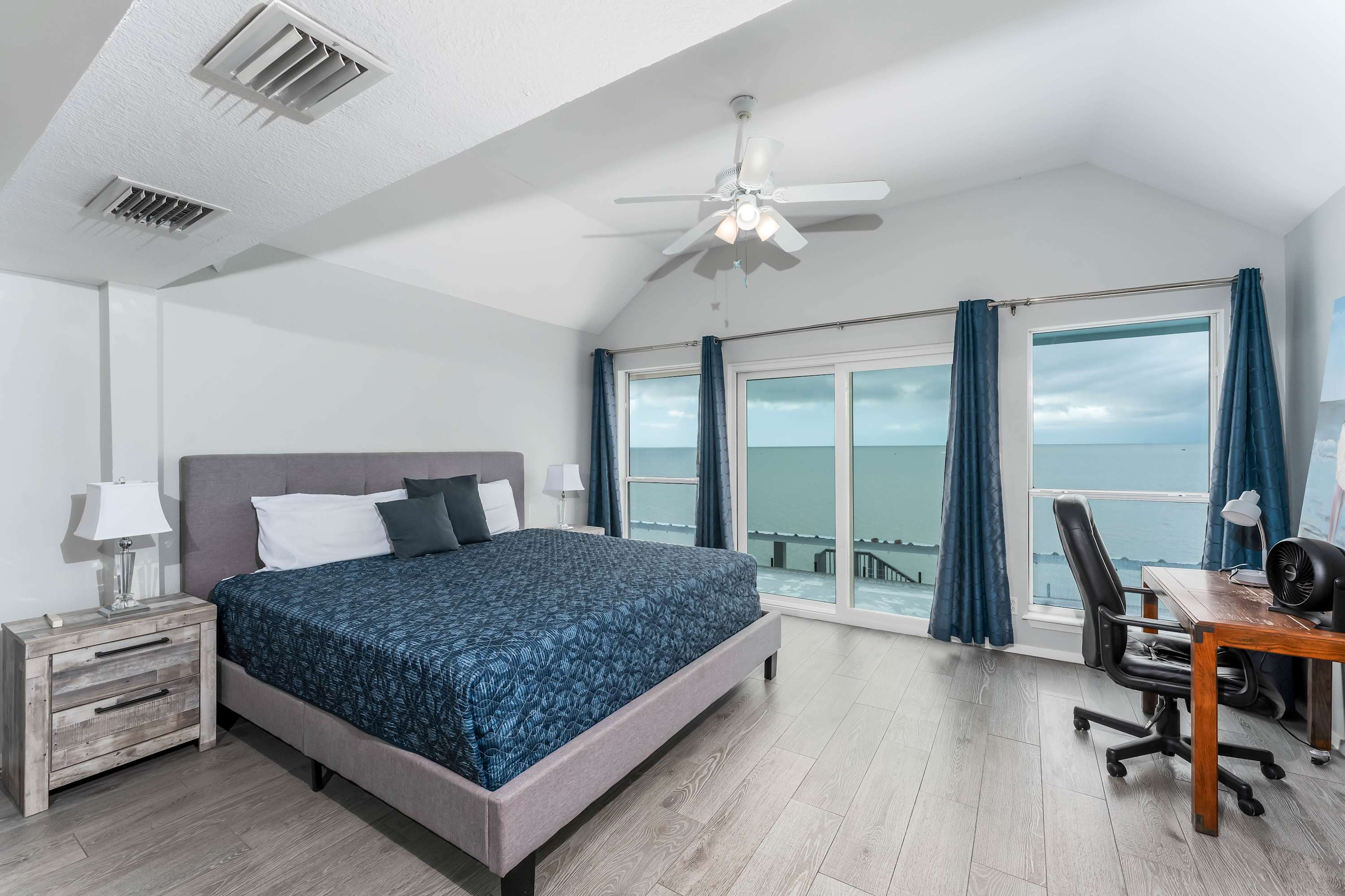 Spacious master bedroom with a king-size bed featuring a blue patterned comforter, light gray walls, and light wood-look flooring, offering panoramic ocean views through large windows and a glass door.