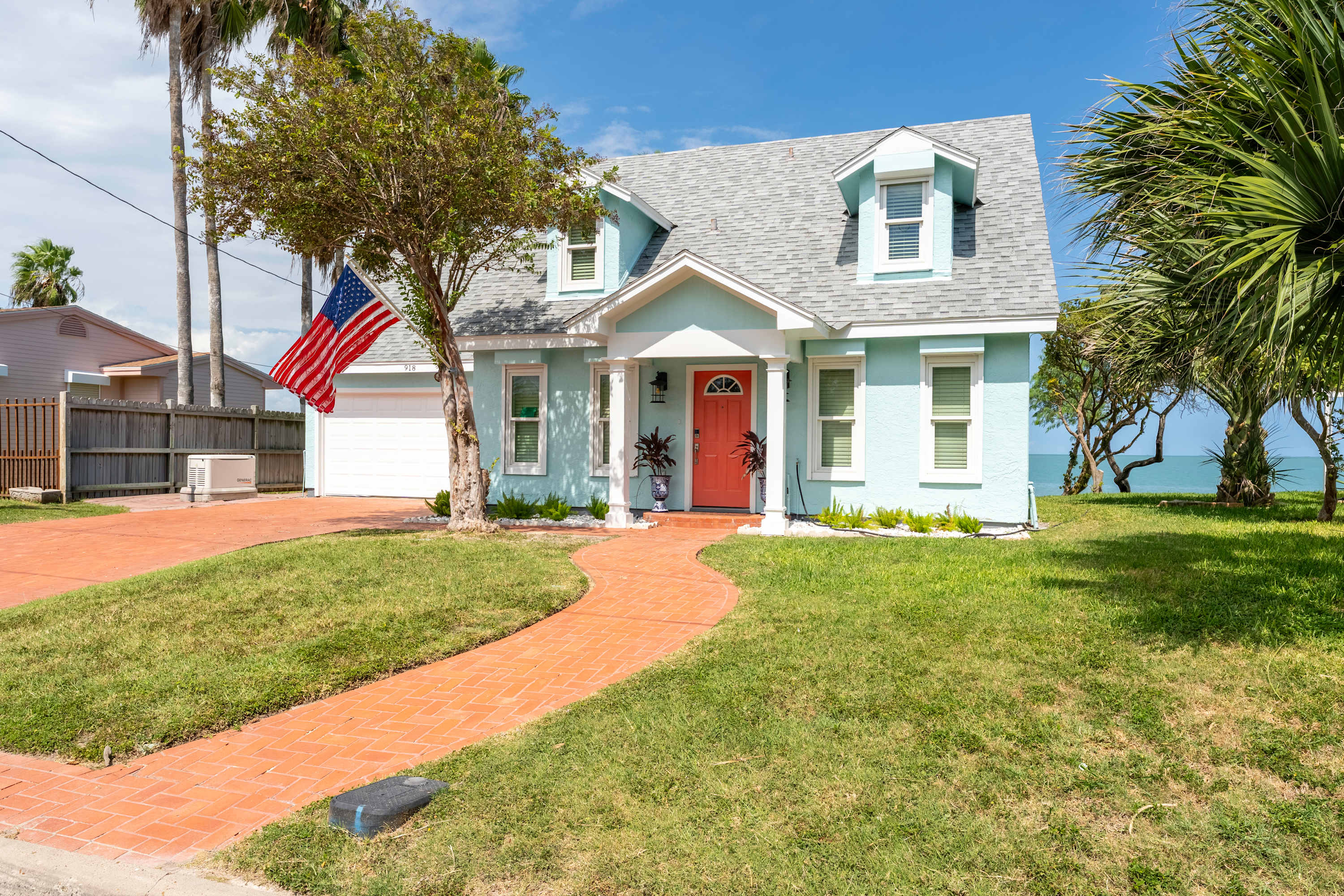 The charming front exterior of a light blue house with a vibrant red front door, a winding brick pathway, and a well-maintained green lawn. An American flag hangs proudly near the garage, with palm trees and the bay visible in the background.
