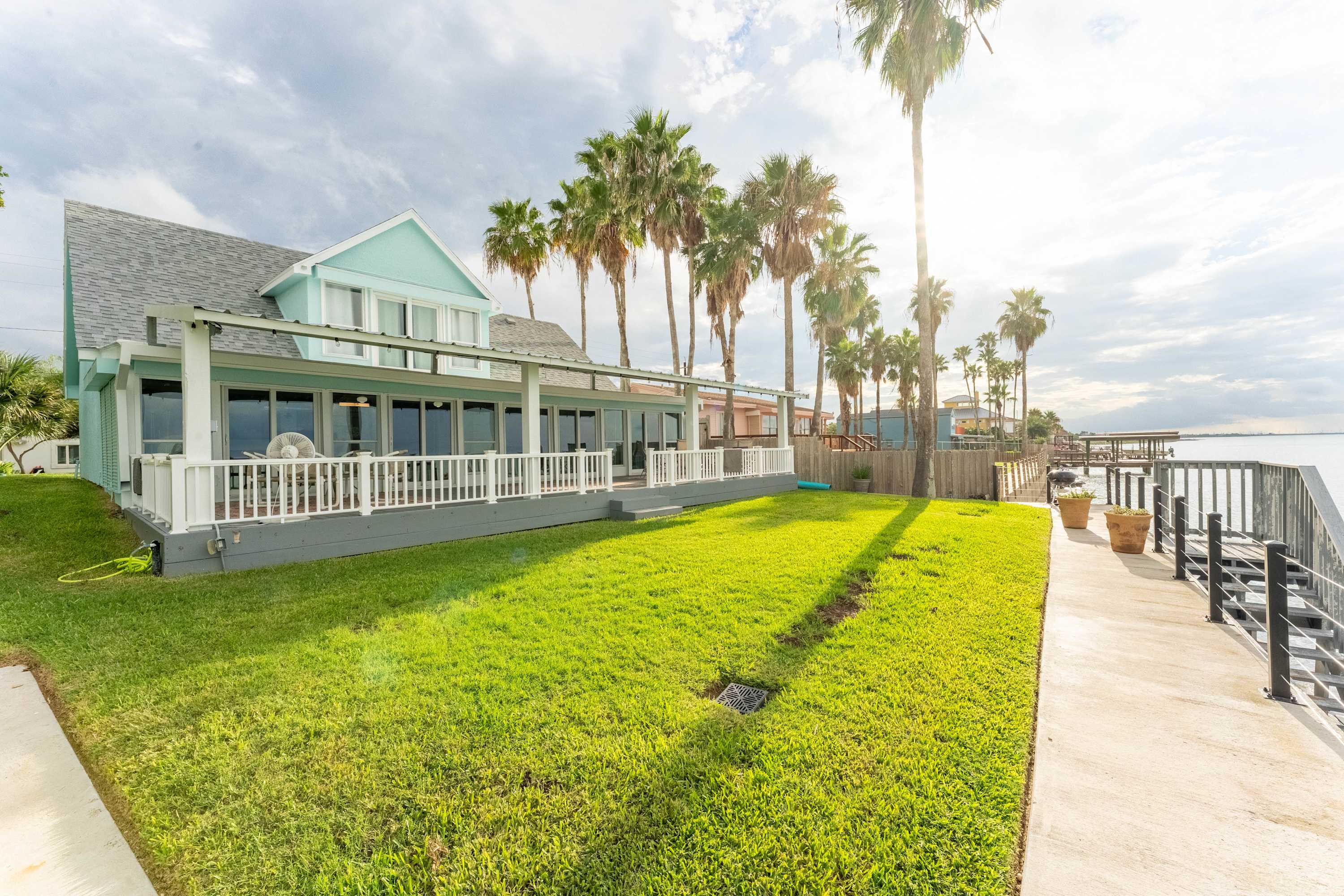 The back exterior of a light blue and white house with a long covered deck, facing a large green lawn and the bay. Tall palm trees line the property, and a concrete walkway leads to a dock.