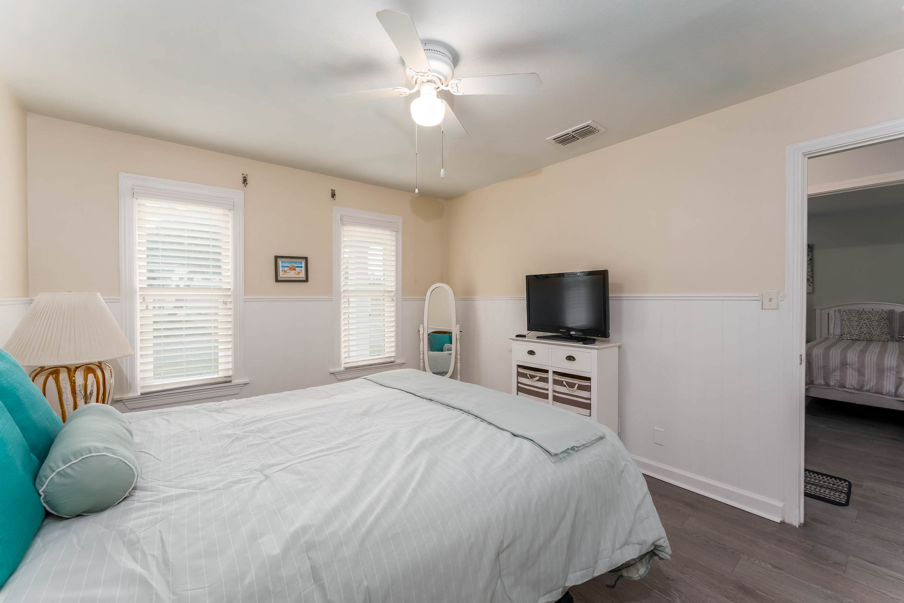 Bedroom with a queen-size bed, light gray wood-look flooring, and white wainscoting, showing a full-length mirror and a white dresser with a TV.