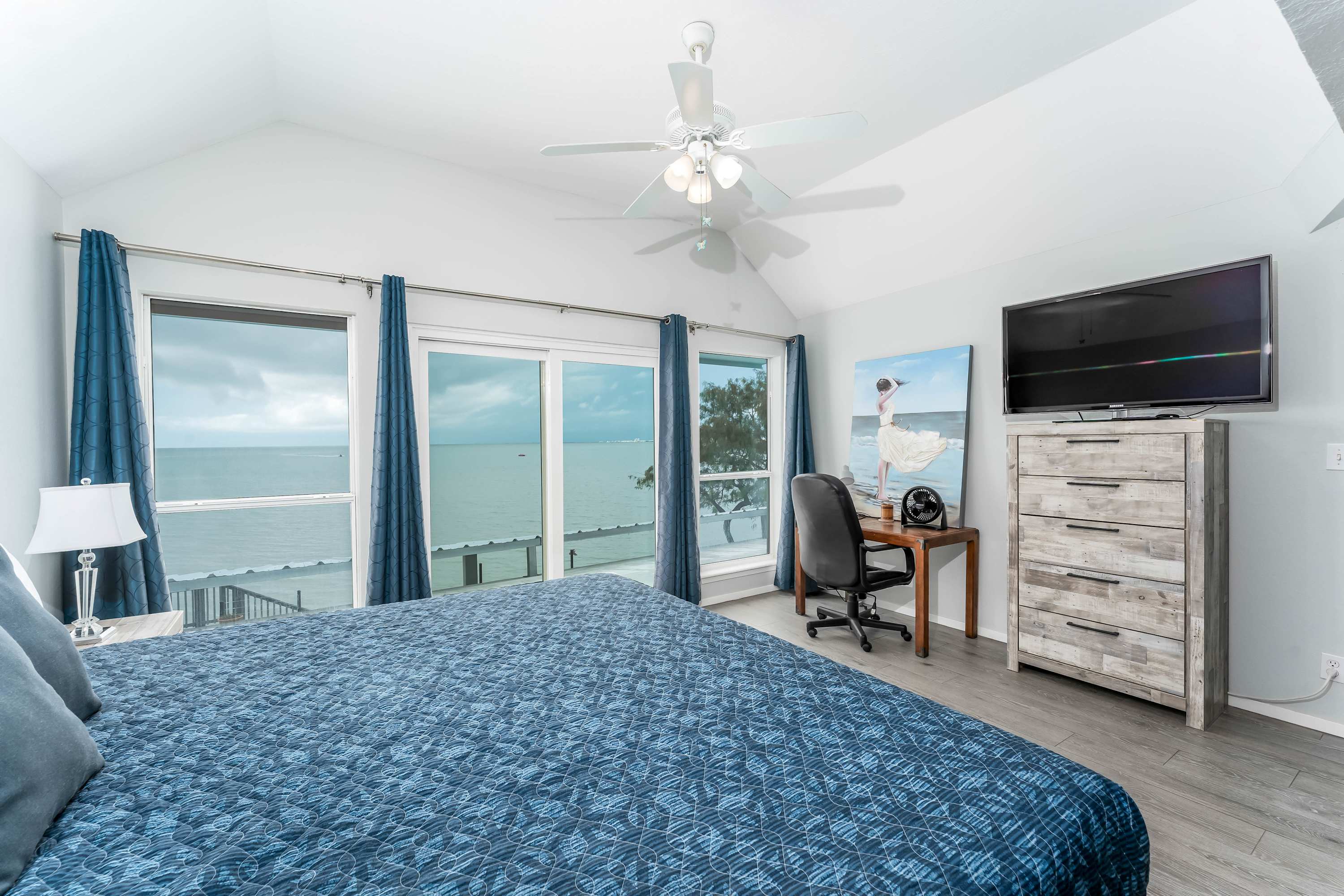 Master bedroom with a king-size bed, light wood-look flooring, and large windows overlooking the ocean, featuring a wooden desk with an office chair and a light wood dresser with a mounted TV.