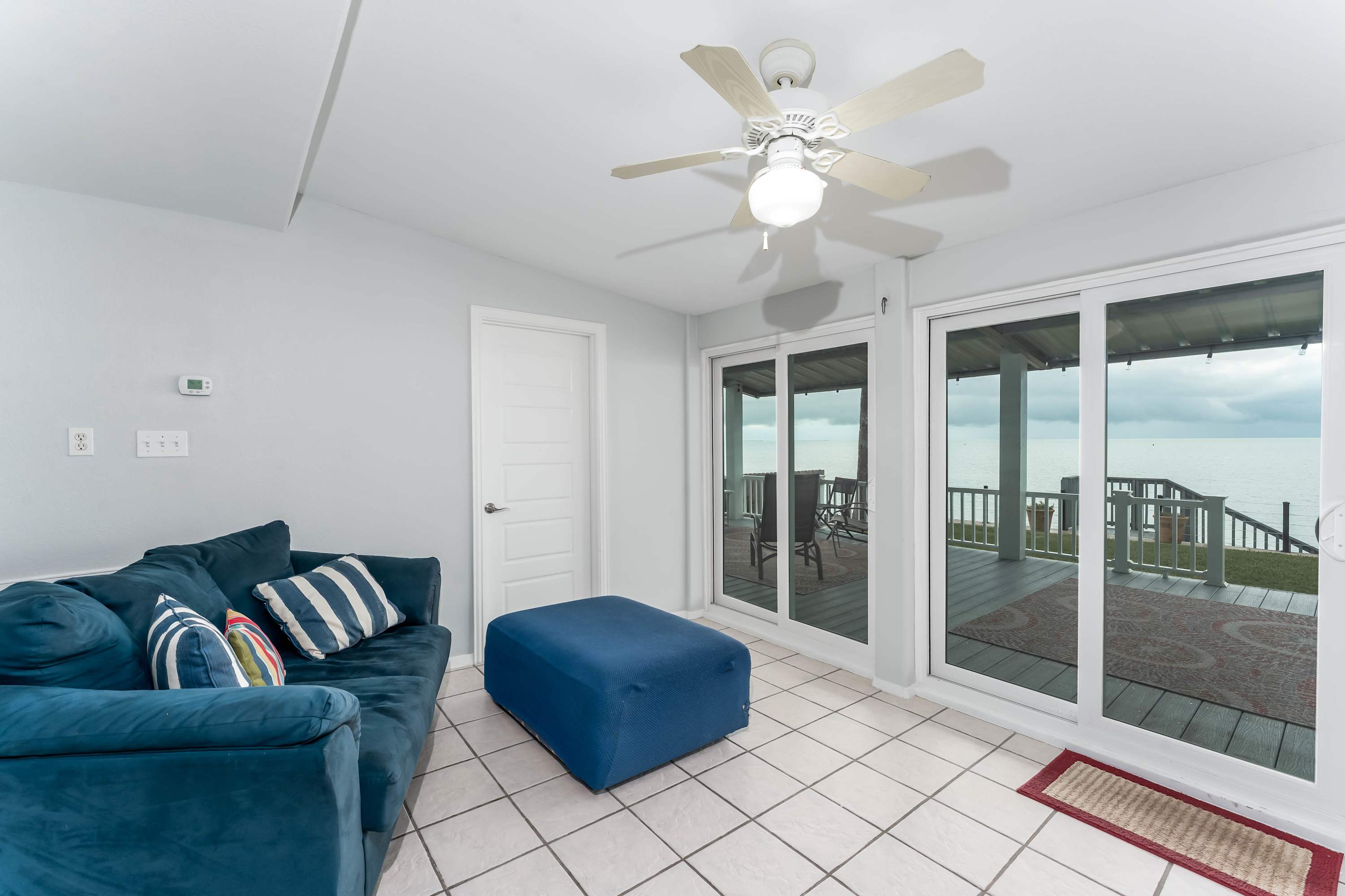 A cozy living area with light blue walls and white tile flooring, featuring a dark blue sectional sofa with striped throw pillows and a matching blue ottoman. Large sliding glass doors offer a view of the outdoor deck and the bay.