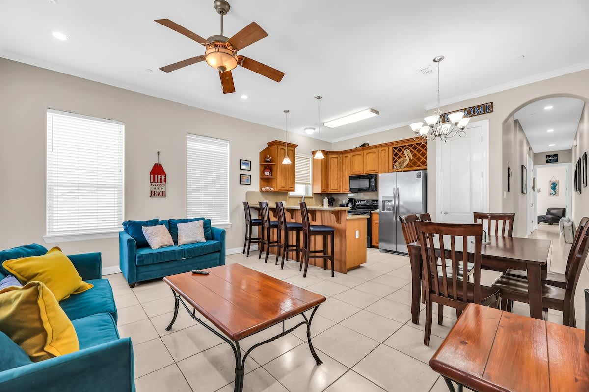 Wide view of the open-concept living area with a teal sofa, a wooden coffee table, and a dining table, extending towards a kitchen equipped with light wood cabinetry and stainless steel appliances, all on light tile flooring.