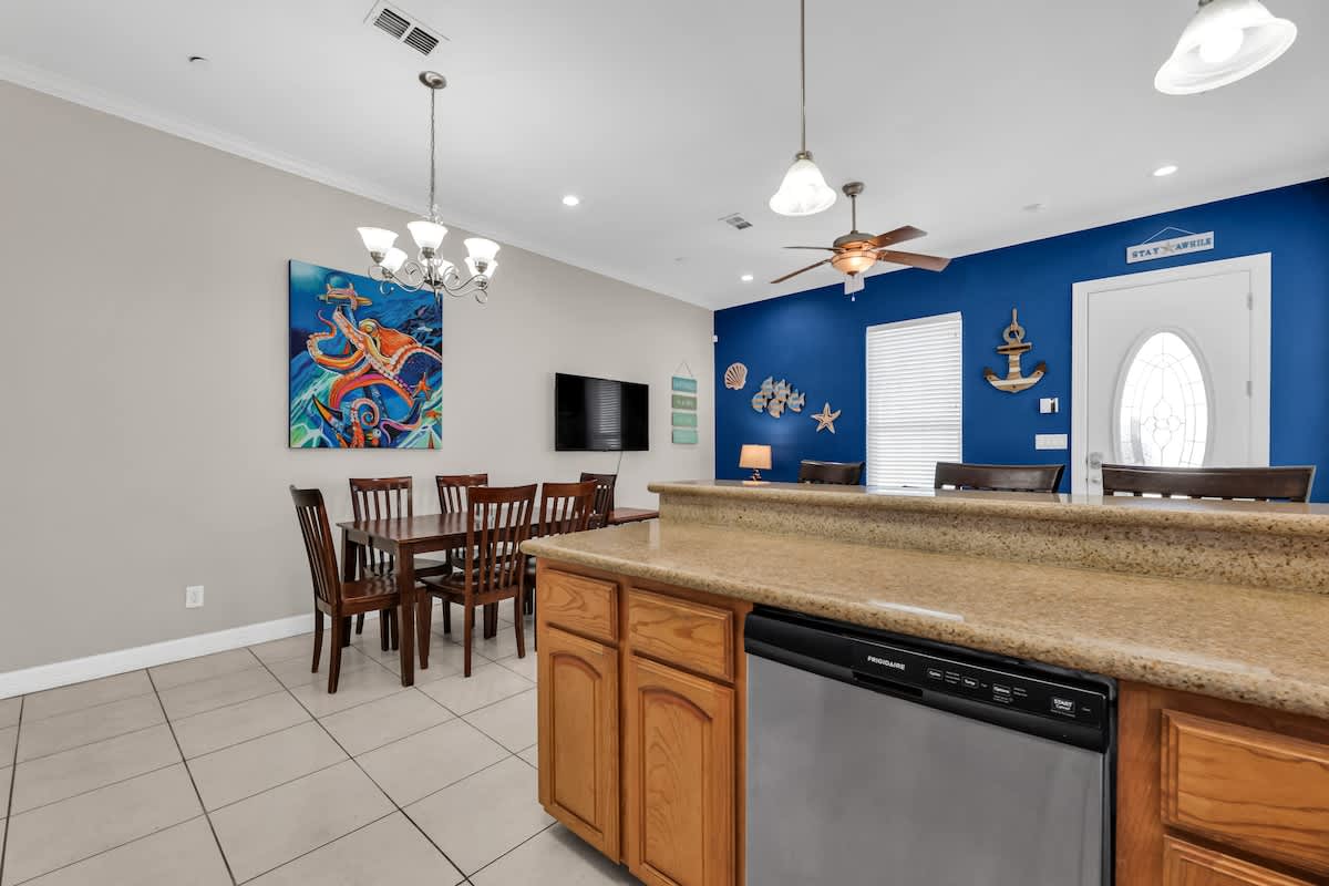 View from the kitchen island looking towards the dining area with a dark wood table and a living room featuring a blue accent wall and teal sofas, all under a ceiling fan and chandelier, with nautical decor throughout.