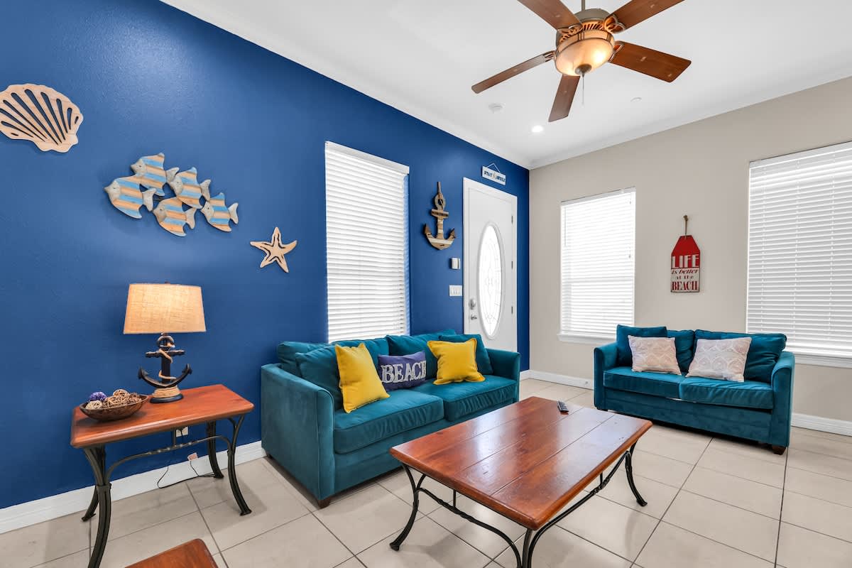Living room with a vibrant blue accent wall adorned with various nautical decor pieces, two teal sofas with yellow and white throw pillows, a wooden coffee table, and white blinds on the windows, all on light tile flooring.