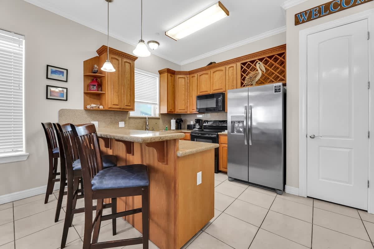 Detailed view of the kitchen featuring light wood cabinetry, speckled granite countertops, a stainless steel refrigerator, a black microwave, and a breakfast bar with dark wood stools, illuminated by pendant lights.