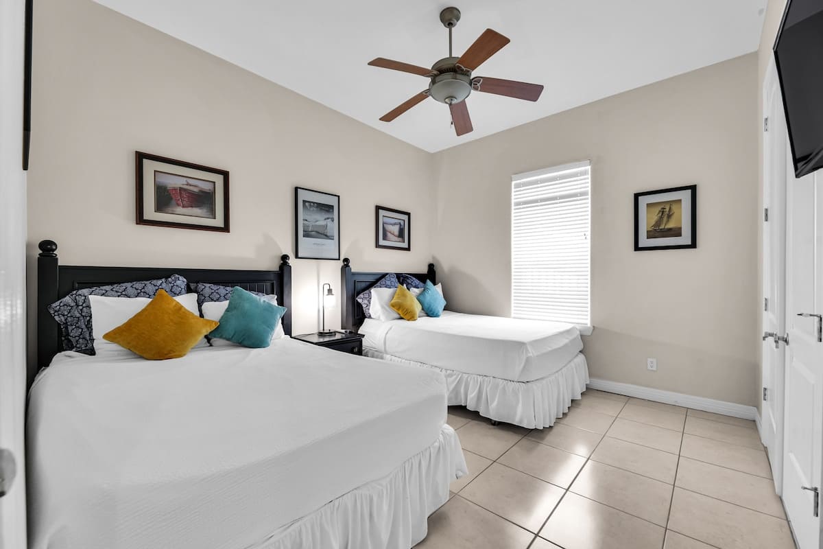 View of the second bedroom showing two beds with black headboards, white bedding, and vibrant yellow and blue pillows, alongside a window with blinds and a wall-mounted TV.