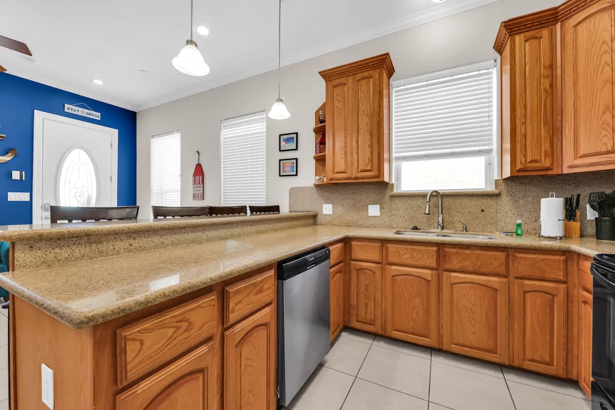 Kitchen counter area with light wood cabinets, speckled granite countertops, a stainless steel dishwasher, a sink positioned under a window with white blinds, and ample counter space, all on light tile flooring.