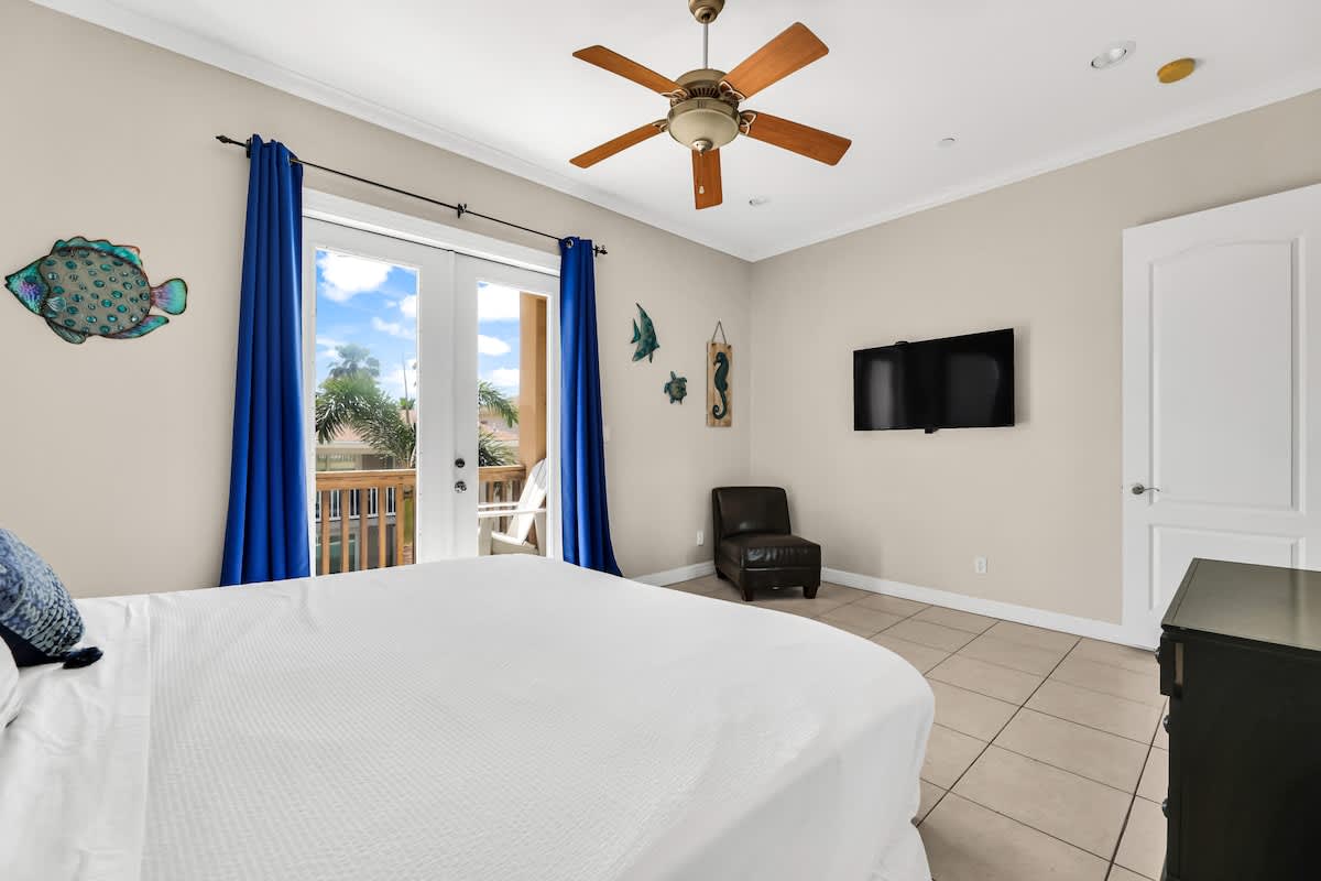 Bright bedroom featuring a large bed with white bedding, a wall-mounted TV, and blue curtains framing glass doors that open to a private balcony with outdoor seating.