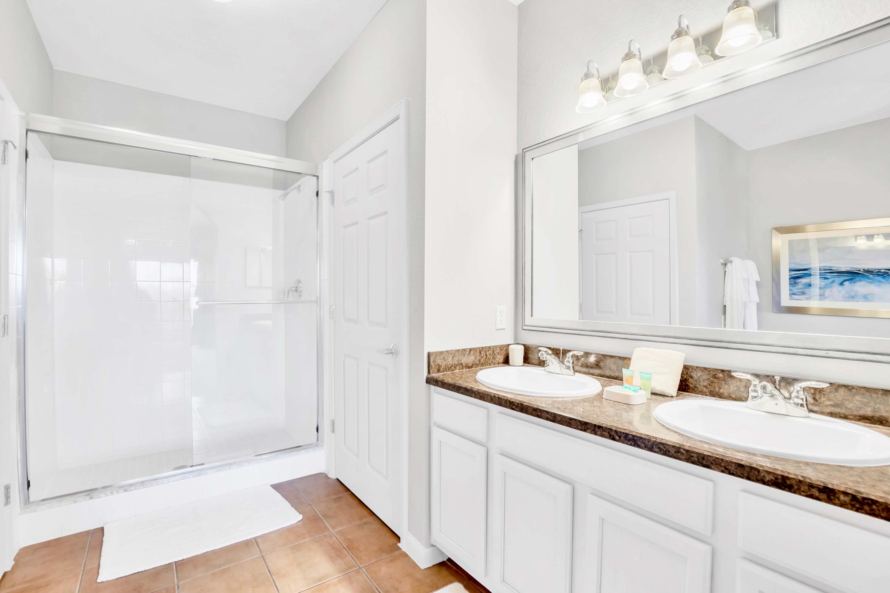 Bright bathroom featuring a double vanity with white sinks and a dark speckled countertop, a large mirror, and a spacious walk-in shower with glass doors. The floor is tiled in a warm brown hue.