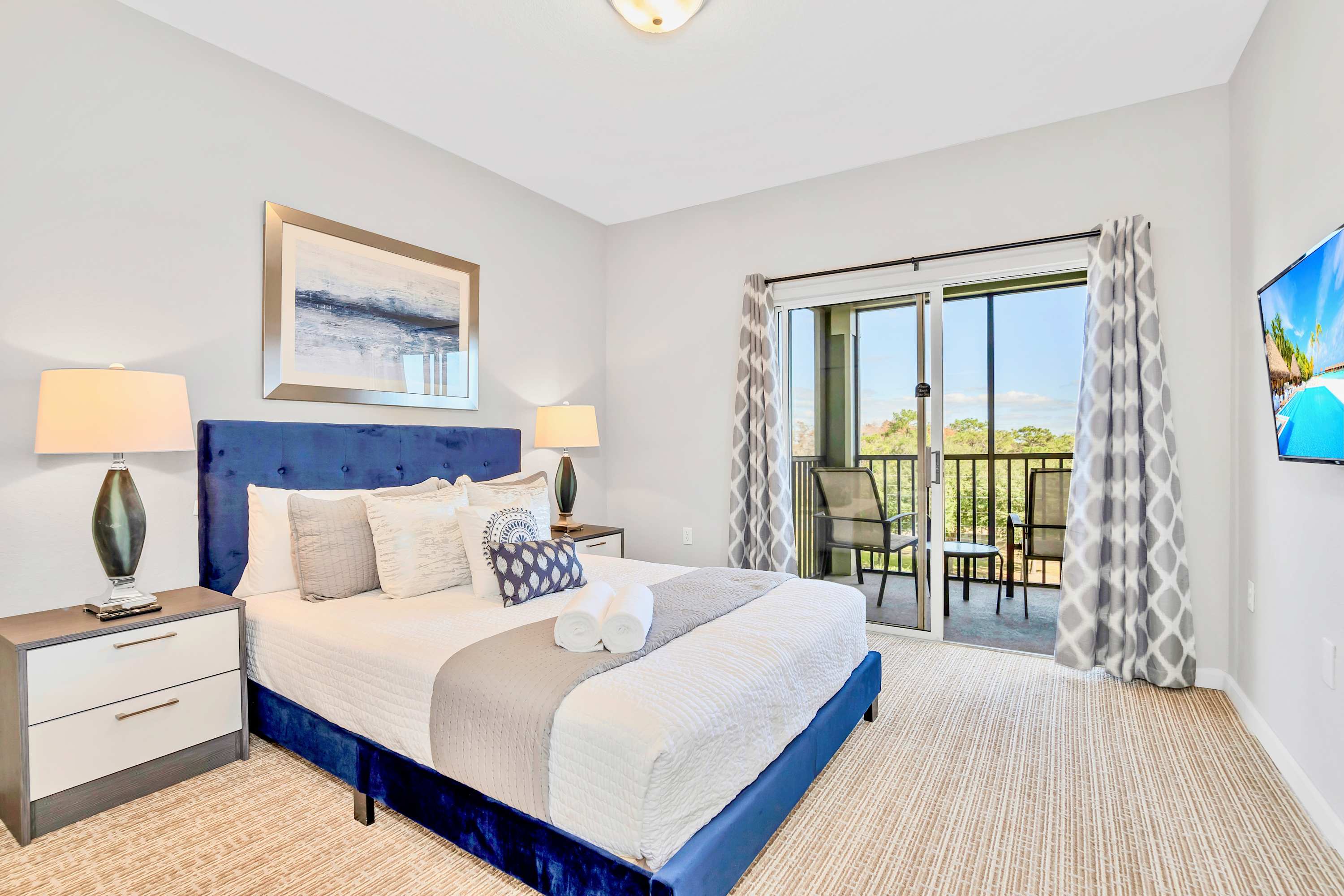 Elegant bedroom with a royal blue velvet bed frame, white and gray bedding, and two bedside tables with lamps. A sliding glass door opens to a private balcony, and a flat-screen TV is mounted on the wall.