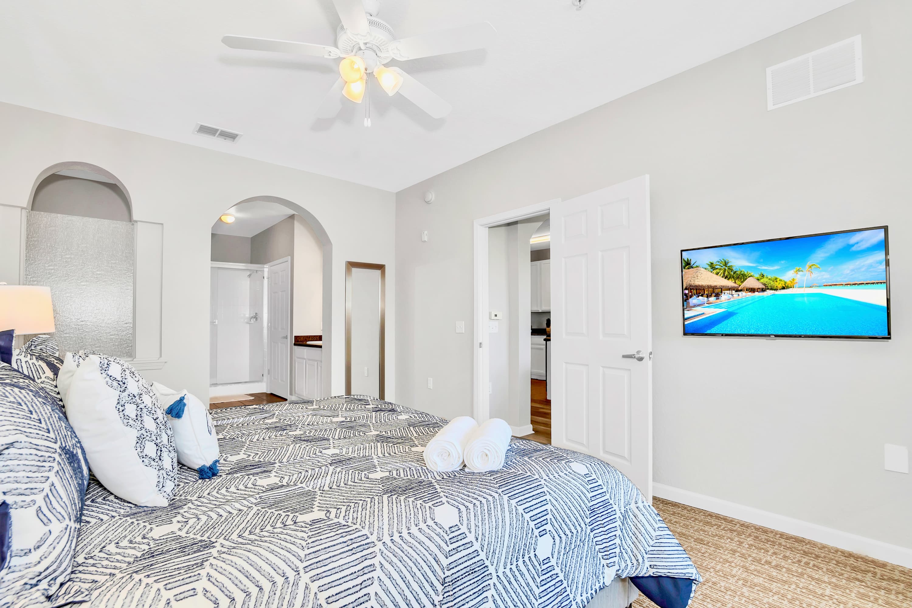 View of the master bedroom with a king-sized bed and a wall-mounted flat-screen TV. An open doorway leads to an adjacent bathroom, revealing a glimpse of a bathtub.