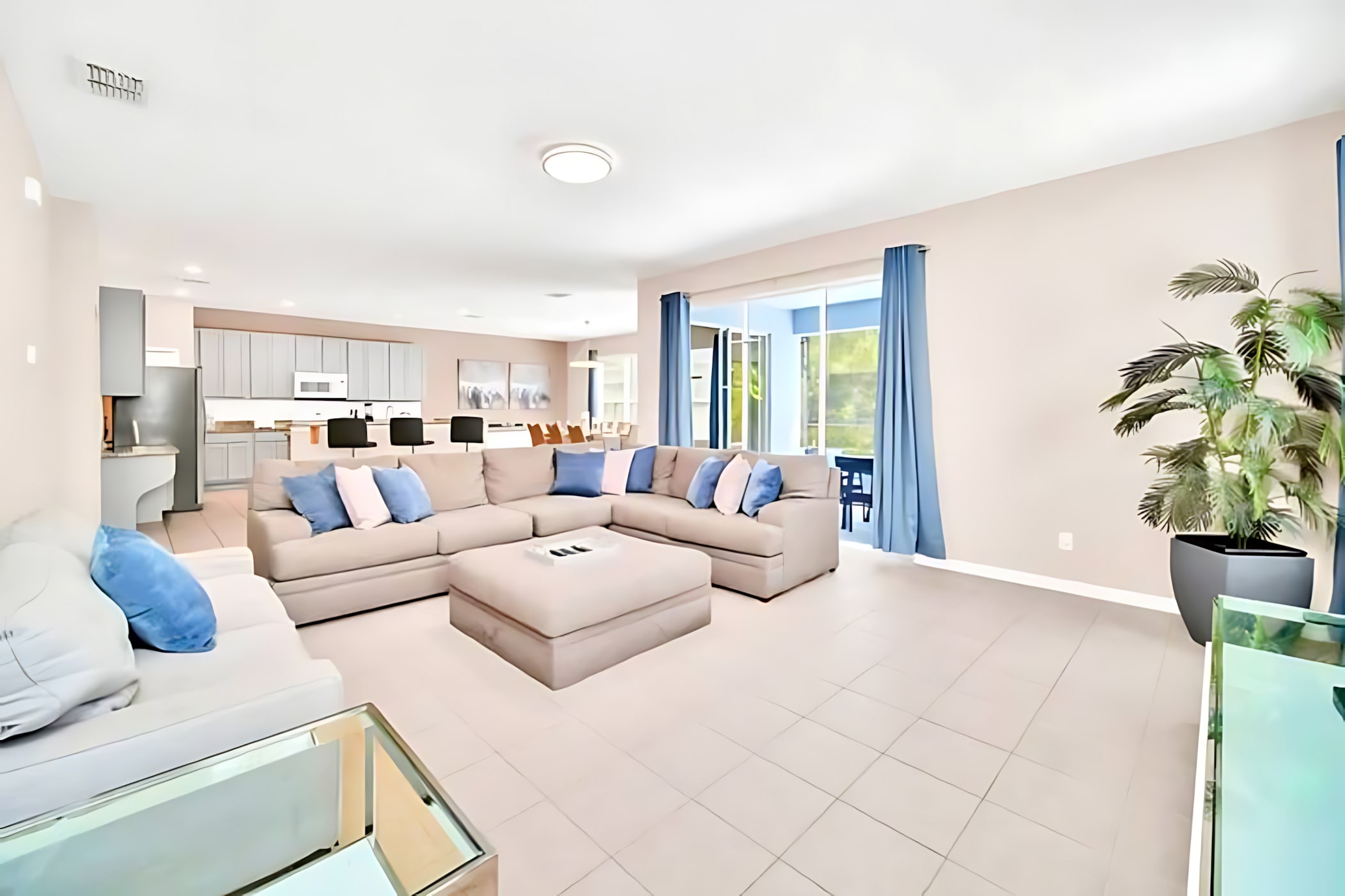 Bright living room with light beige tiled floors, a large L-shaped sectional sofa with blue and white throw pillows, a matching ottoman, and blue curtains framing sliding glass doors leading to an outdoor area.