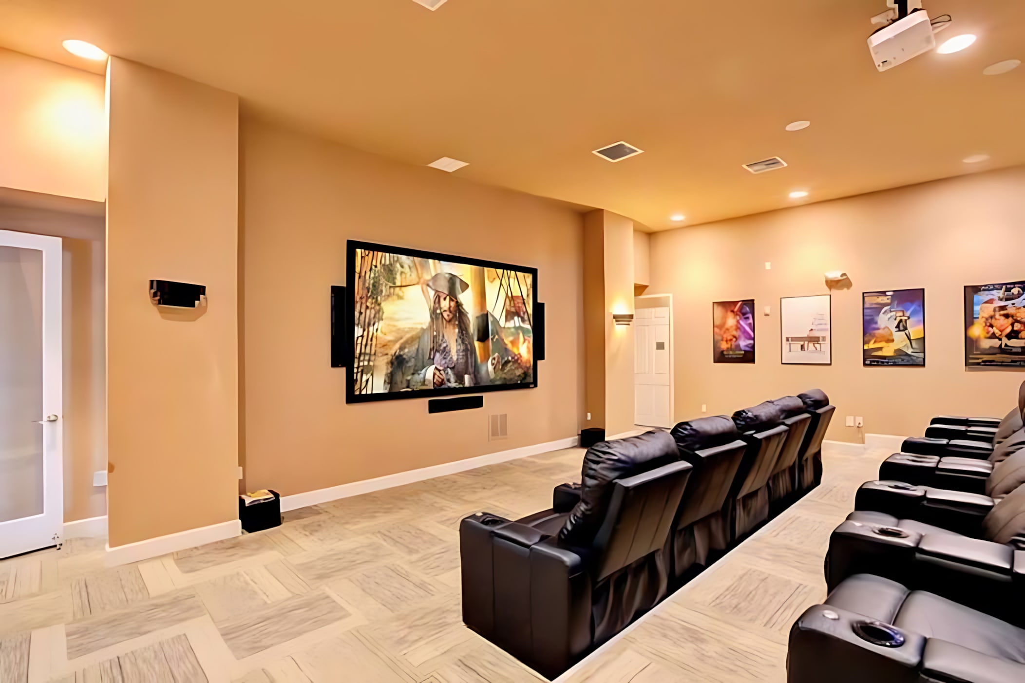 A dedicated home theater features multiple rows of black leather recliners facing a large wall-mounted screen displaying a movie. The room has light beige walls, recessed lighting, and movie posters on the side wall.