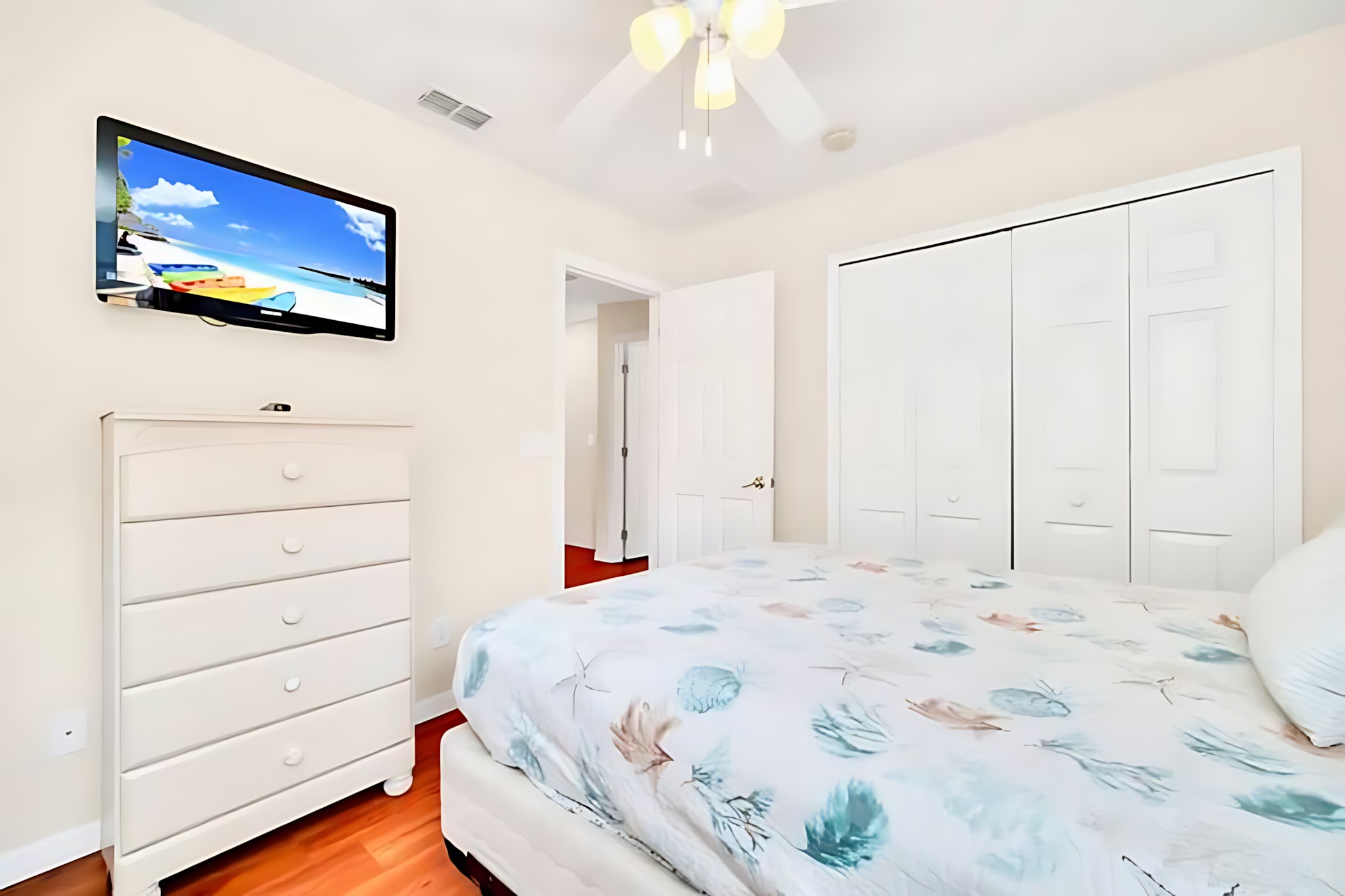 This view of the bedroom shows a wall-mounted flat-screen TV above a white dresser, a queen-size bed with coastal bedding, and white bi-fold closet doors.
