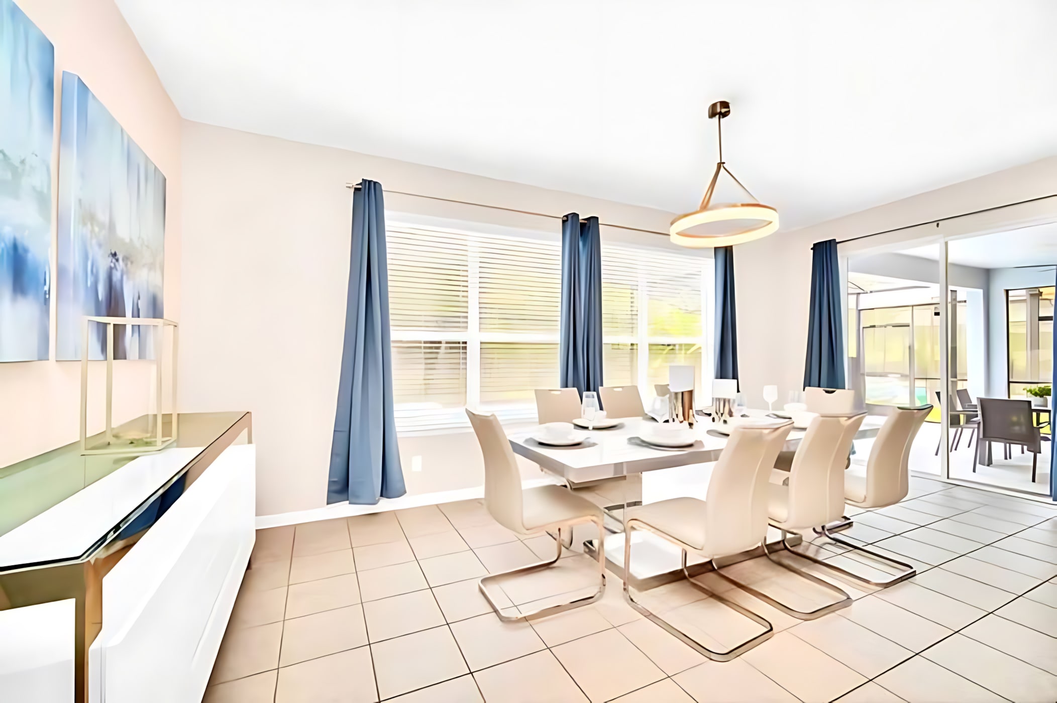 Elegant dining room with light tiled floors, a rectangular white dining table set for six with modern cream chairs, and blue curtains framing a window and a sliding glass door leading to an outdoor area.