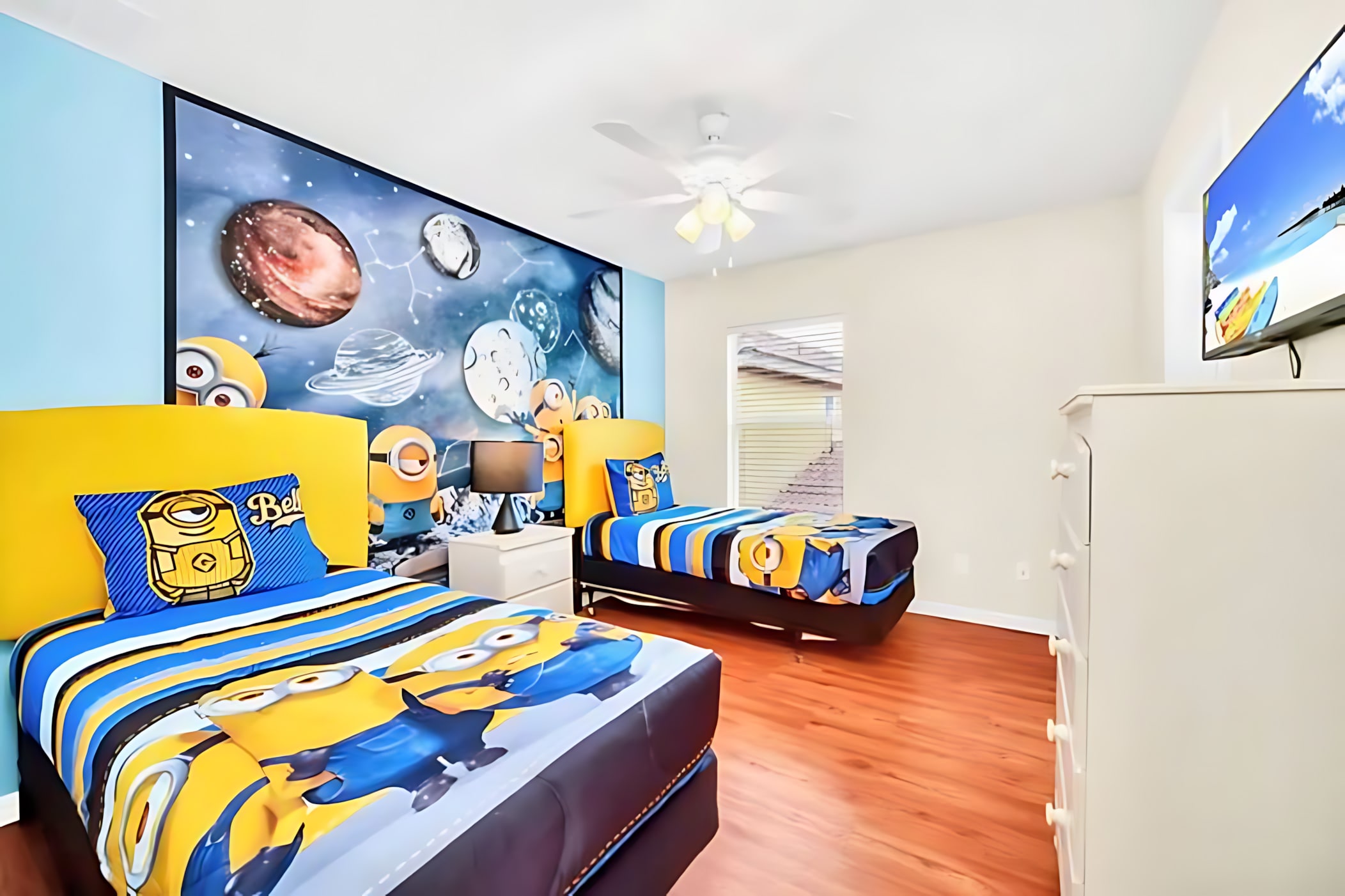 Vibrant Minions-themed bedroom with two twin beds, bright yellow headboards, character bedding, and a large wall mural depicting planets and Minions in space.
