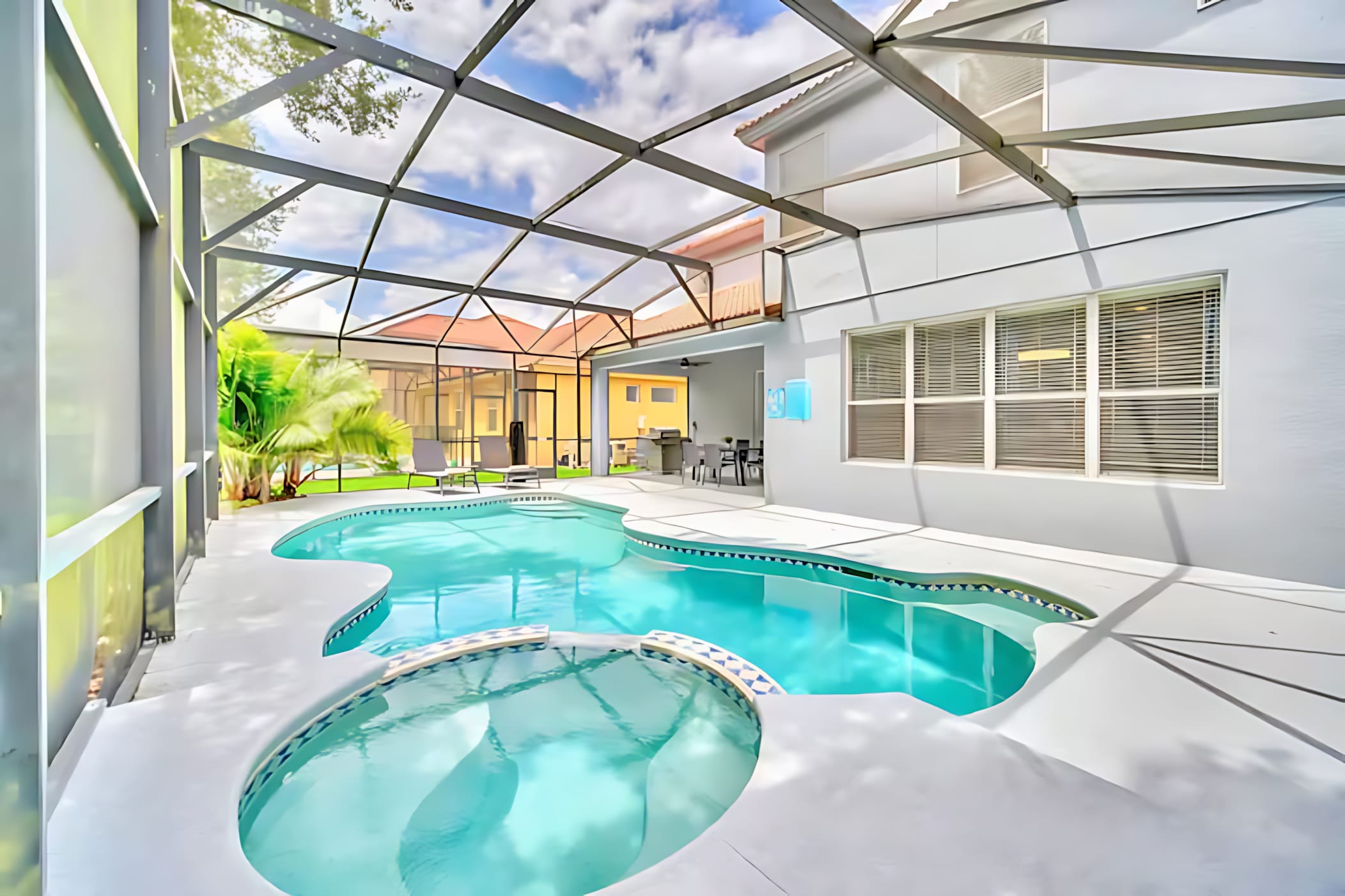 A bright, screened-in outdoor pool area features a freeform pool with an attached hot tub, set against the backdrop of a modern house. The white concrete deck provides ample space for relaxation, with a glimpse of outdoor dining furniture.