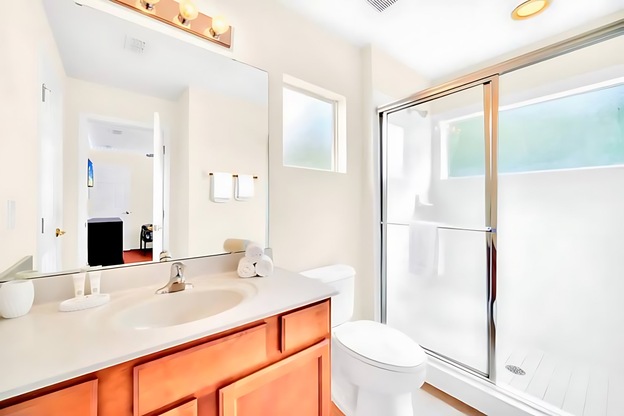 Bright bathroom with a single wood vanity, light countertop, large mirror, and a glass-enclosed walk-in shower, featuring white towels and modern light fixtures.