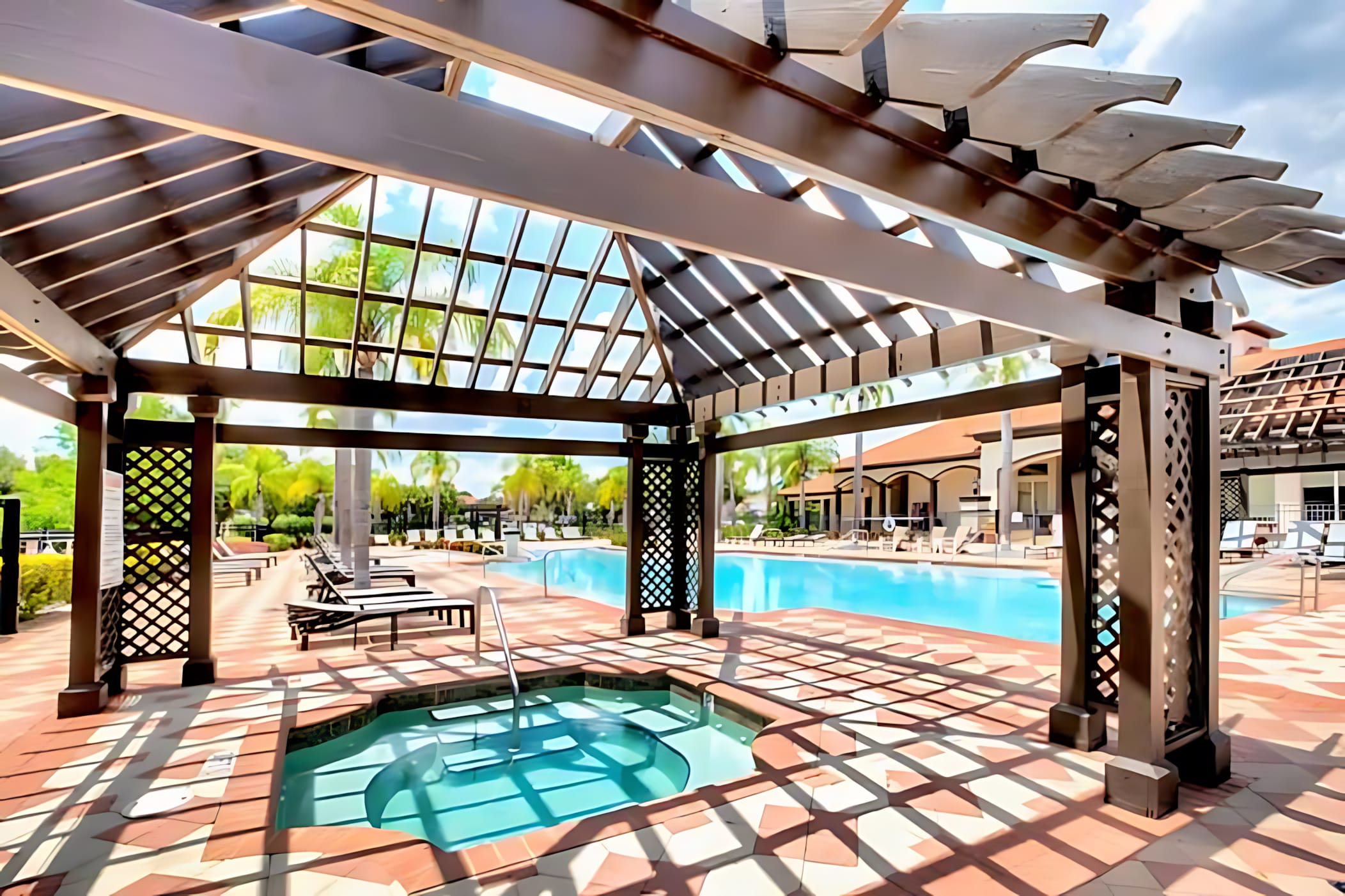 Shaded outdoor hot tub located under a wooden pergola, adjacent to a large swimming pool, with patterned paver decking and palm trees in the background.