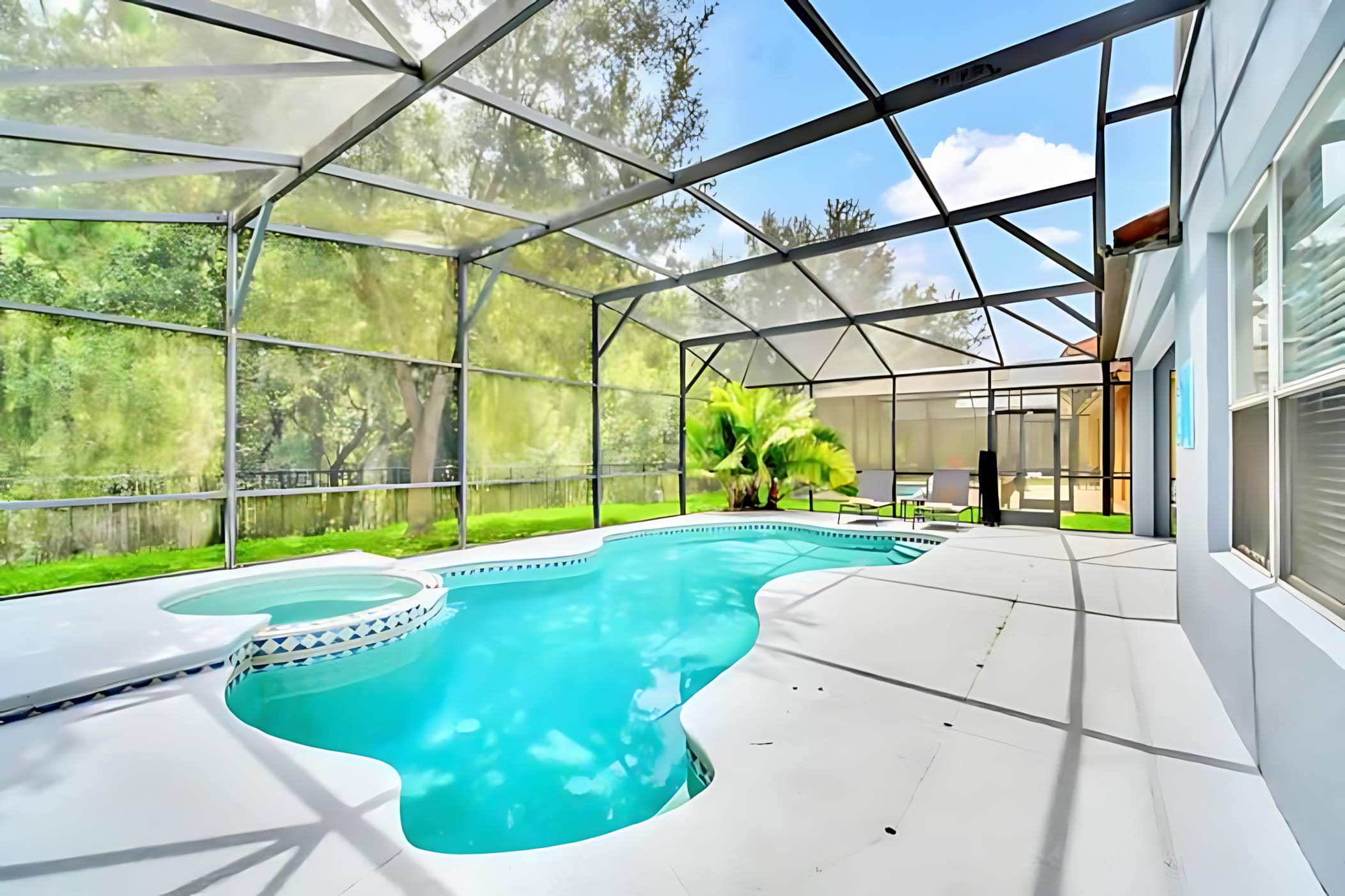 A large, screened-in outdoor pool area features a freeform pool with an integrated hot tub, surrounded by a white concrete deck. Lush green trees and a manicured lawn are visible beyond the screen, offering a private setting.