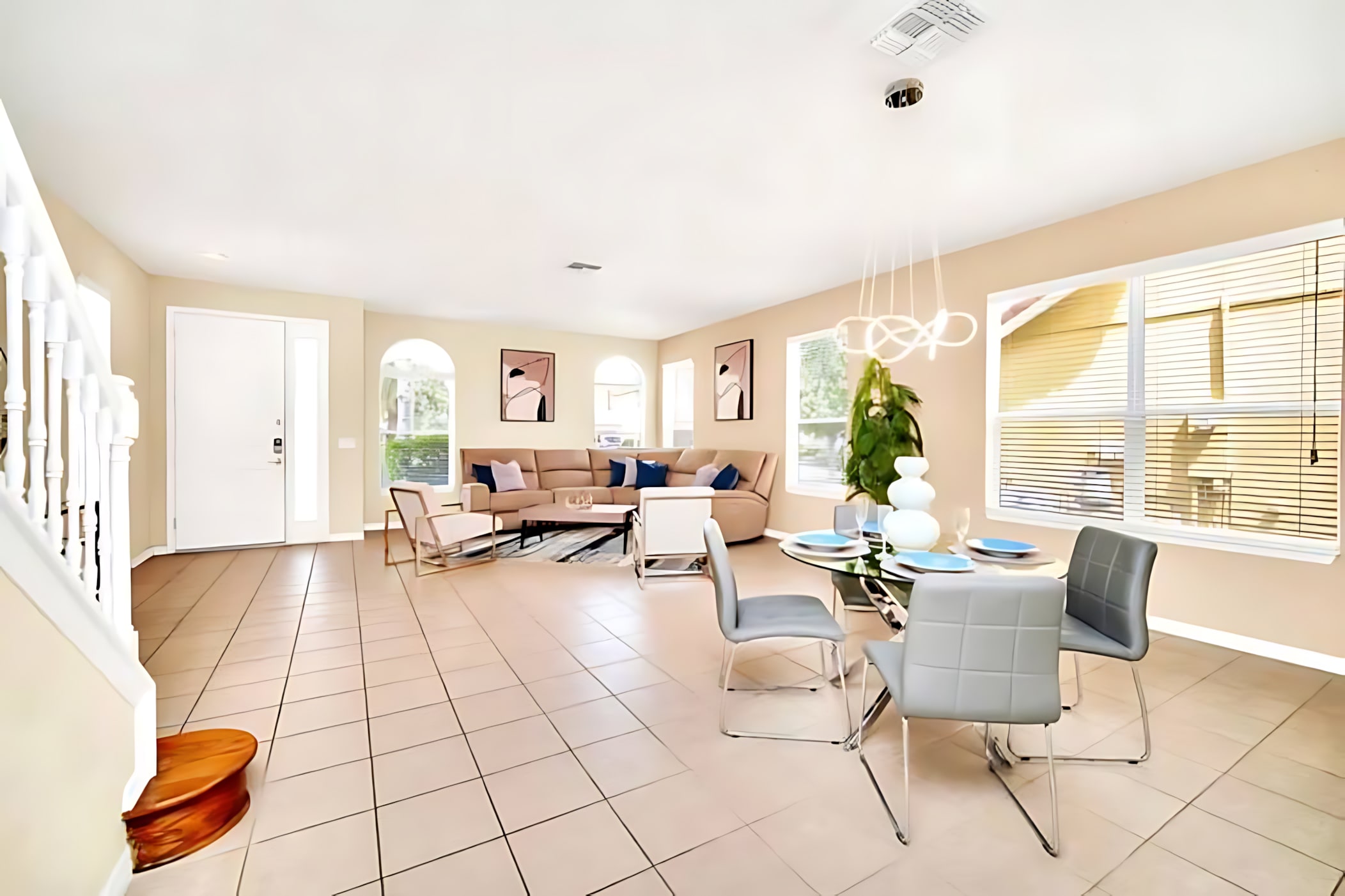 Bright open-plan living and dining area with light tiled floors, a beige sectional sofa with blue pillows, a glass-top coffee table, and a round glass dining table with gray chairs under a modern chandelier.