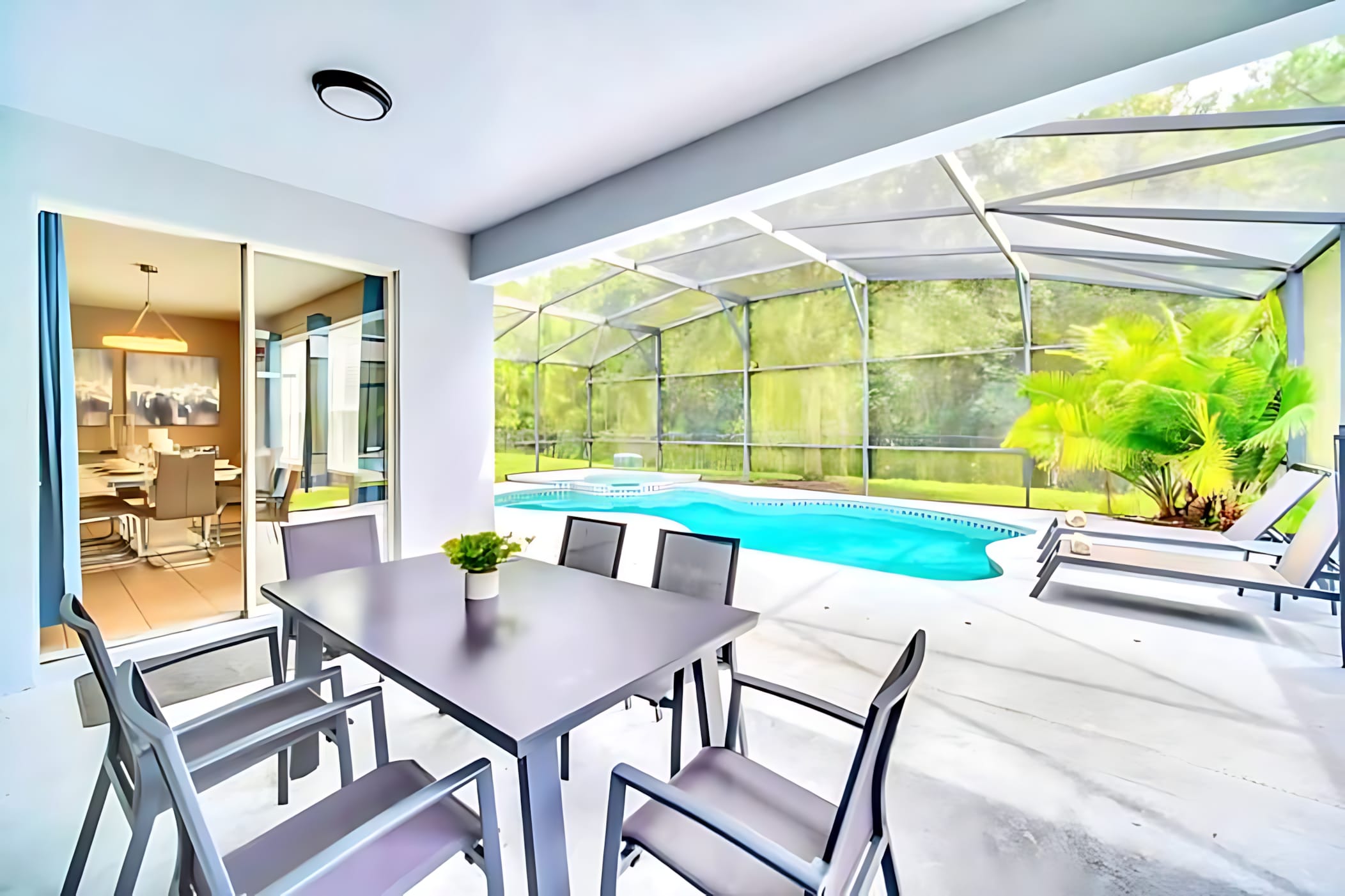 A covered patio area features a modern gray outdoor dining table with six chairs, overlooking a screened-in pool and hot tub. Sliding glass doors connect the patio to the interior dining area, offering seamless indoor-outdoor living.