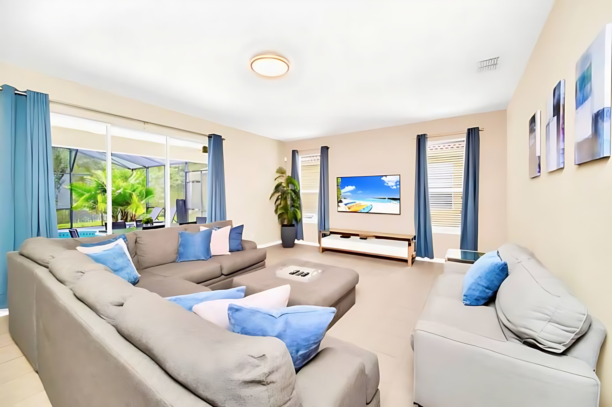 A spacious living room features a large L-shaped gray sectional sofa with blue and white accent pillows, facing a wall-mounted flat-screen TV. Sliding glass doors lead to the outdoor pool area, providing natural light.