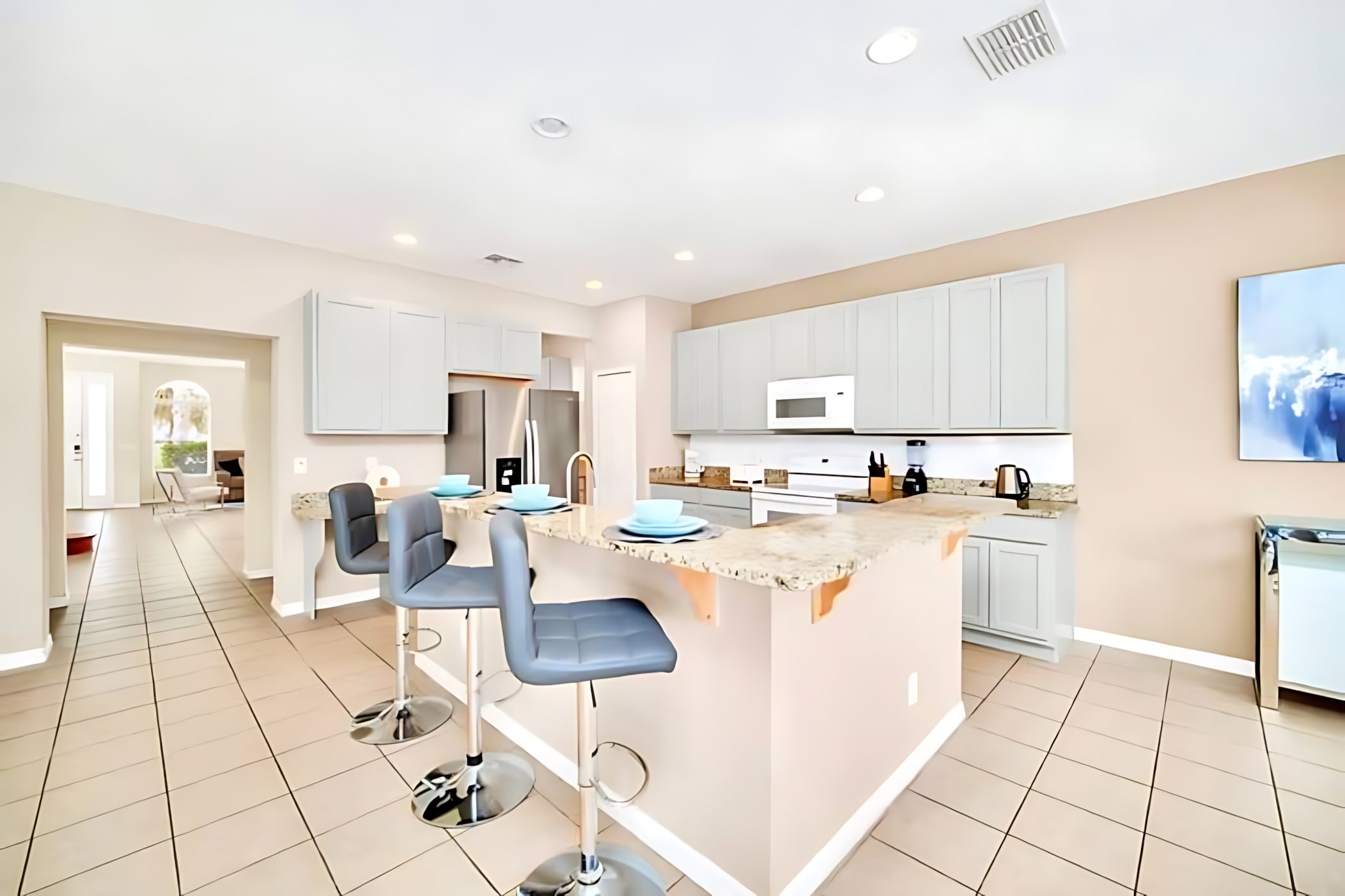Spacious kitchen featuring light tiled floors, light gray cabinets, granite countertops, a large island with three gray bar stools, and stainless steel appliances including a refrigerator and microwave.