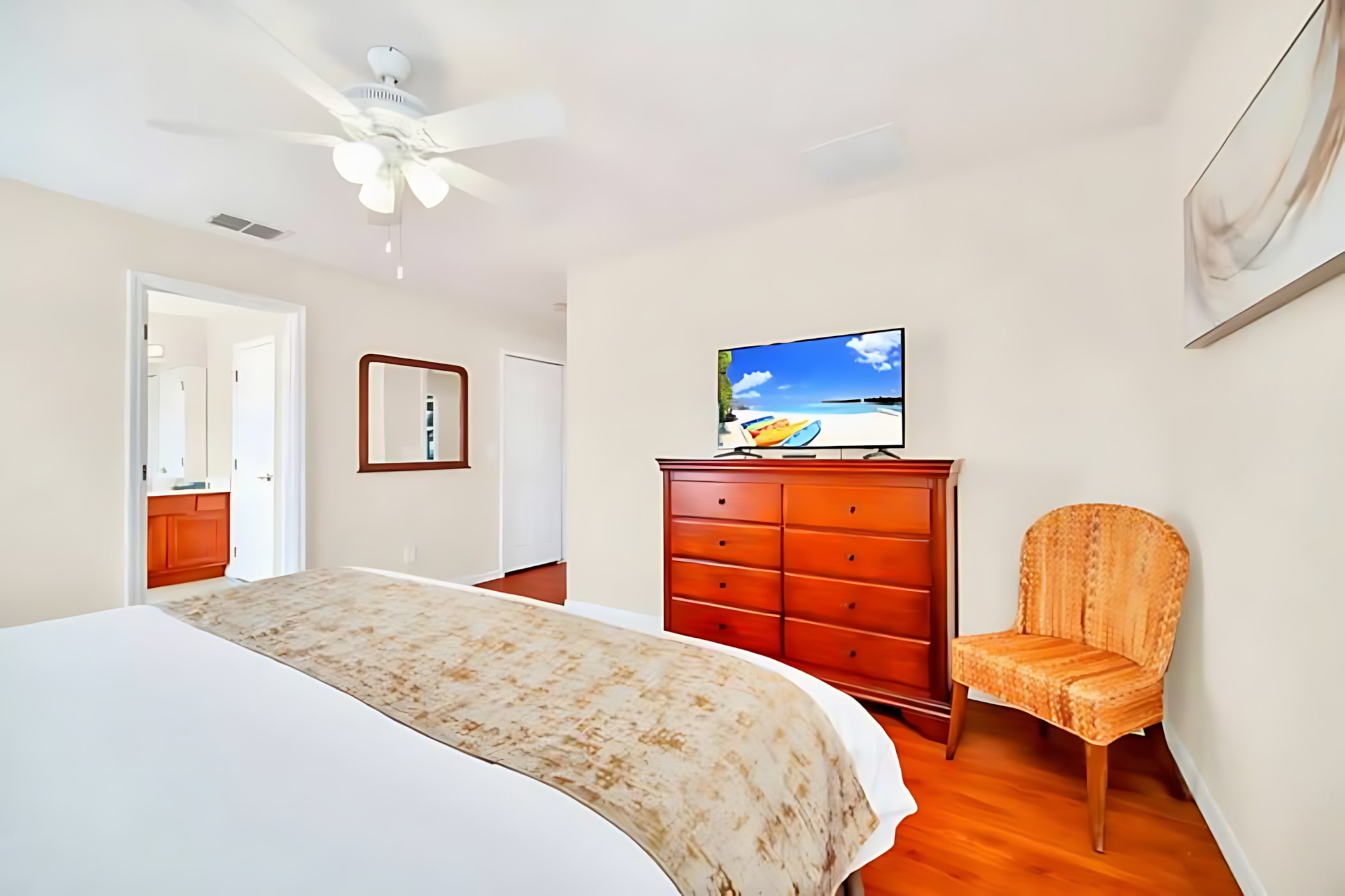 A bright bedroom features a king-size bed with white and gold bedding, a wooden dresser with a flat-screen TV, and a wicker chair, all set on polished hardwood floors. A ceiling fan hangs overhead.