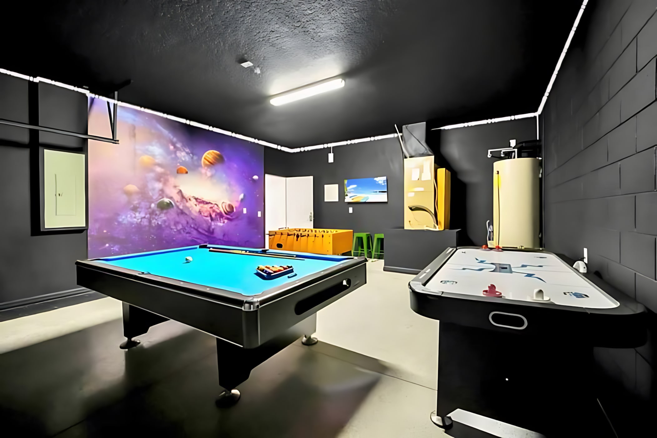 A spacious game room with dark walls and ceiling is equipped with a blue-felt pool table, an air hockey table, and a foosball table. A vibrant space-themed mural and a wall-mounted TV add to the entertainment options.
