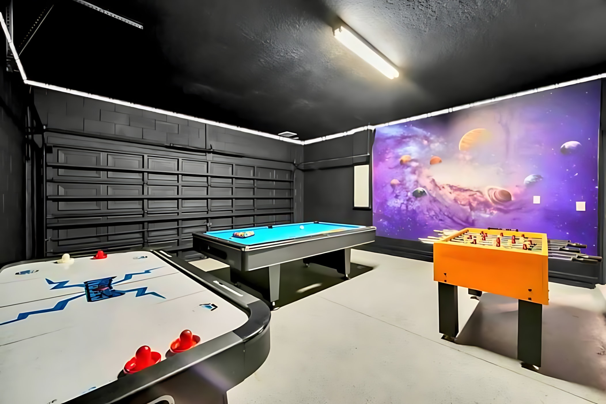 A vibrant game room with dark walls and ceiling features a blue-felt pool table, an air hockey table, and a foosball table. A colorful space-themed mural adorns one wall, adding a playful touch.