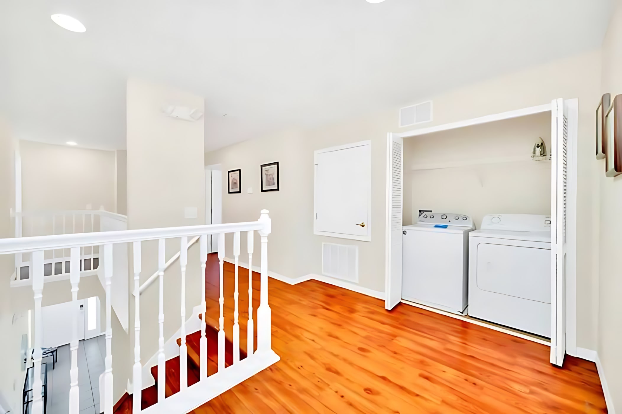 Upstairs landing with polished hardwood floors, white banister, and a bi-fold closet revealing a white top-loading washing machine and dryer, indicating a dedicated laundry area.