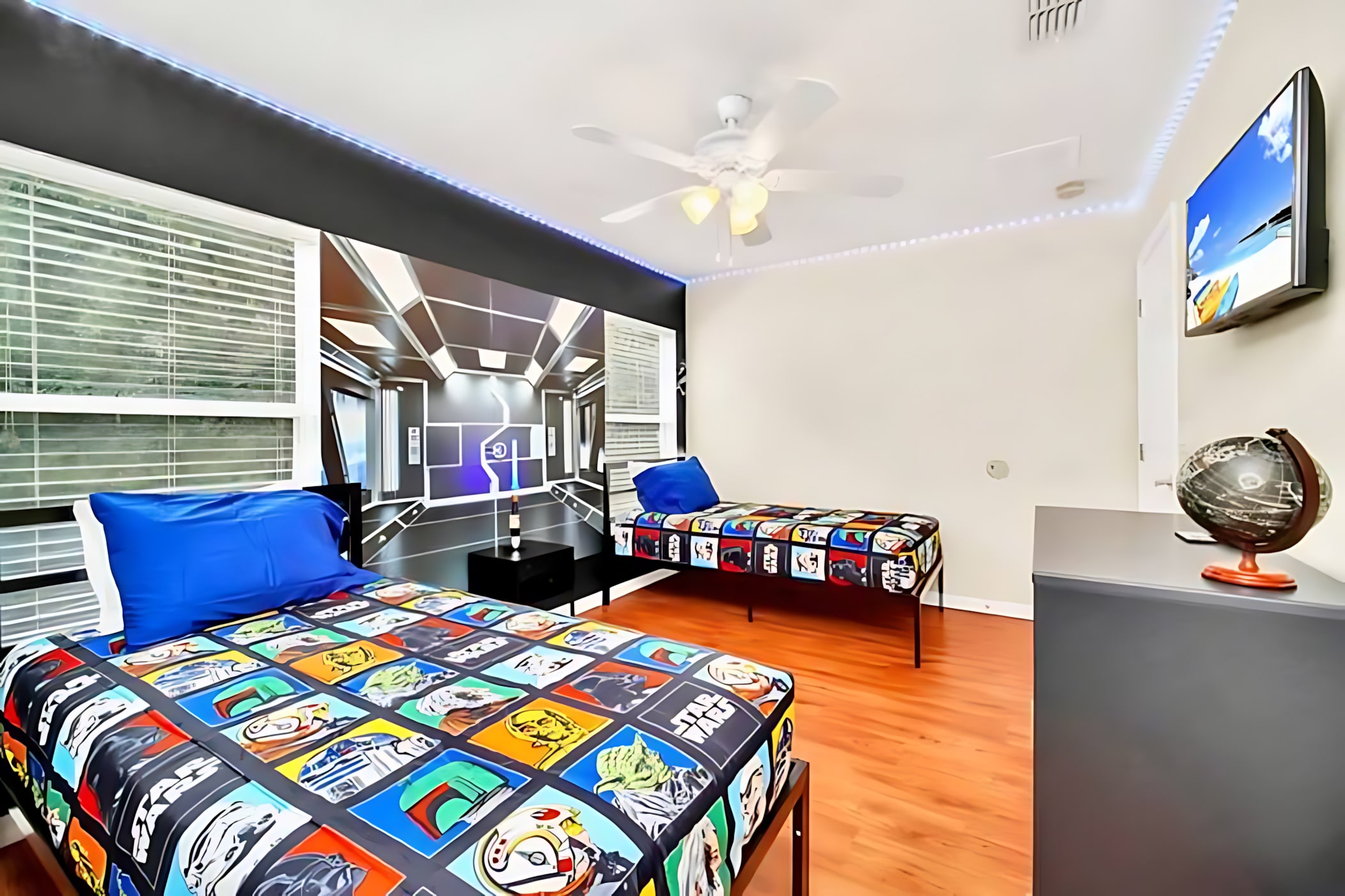 Wide view of the Star Wars themed bedroom with two twin beds, vibrant character bedding, a large wall mural, and a wall-mounted TV, illuminated by ceiling fan and blue LED lights.