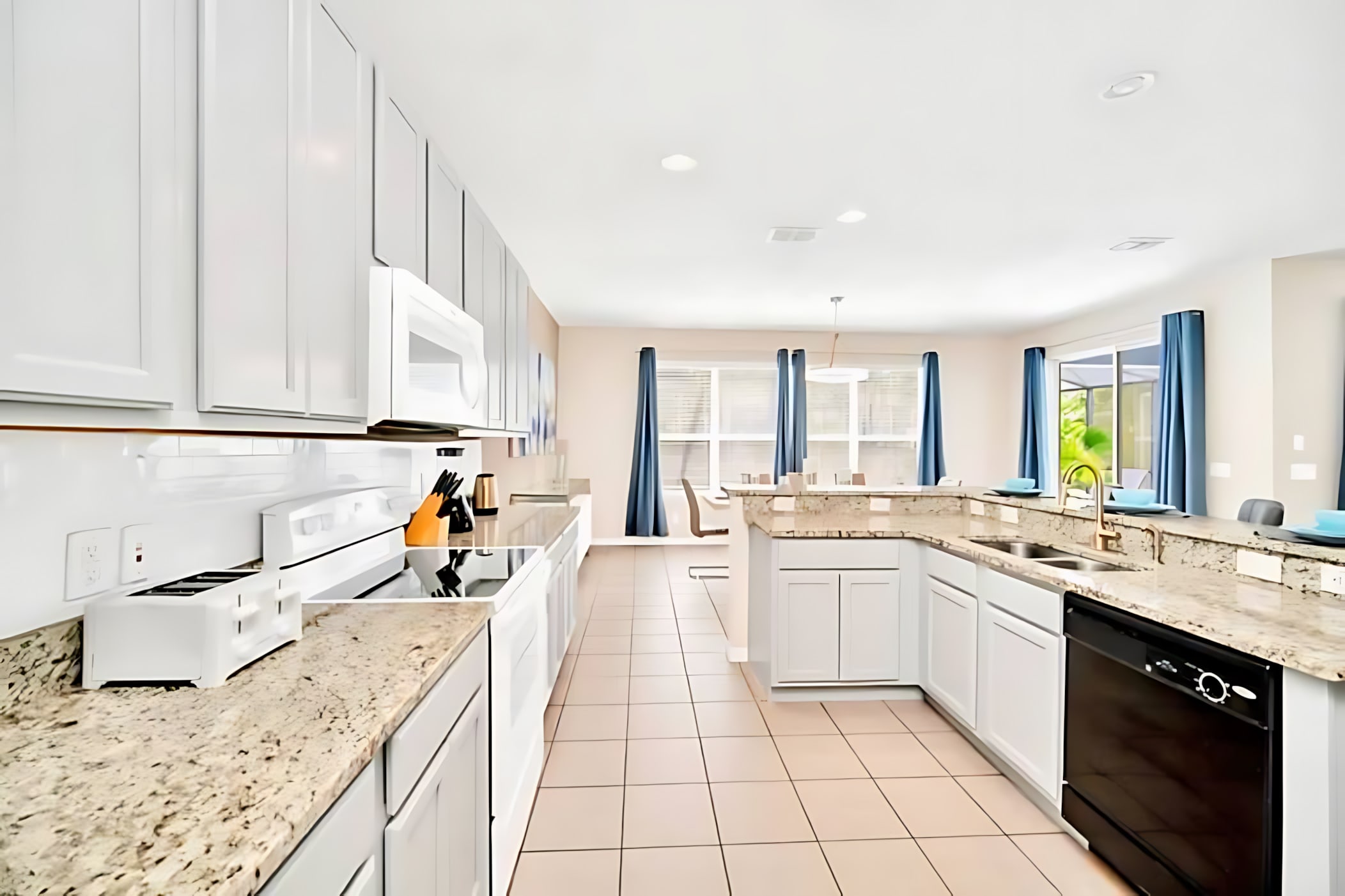 Modern kitchen with light tiled floors, white upper and lower cabinets, light granite countertops, a white electric range, microwave, and a black dishwasher, with a view towards the dining area.