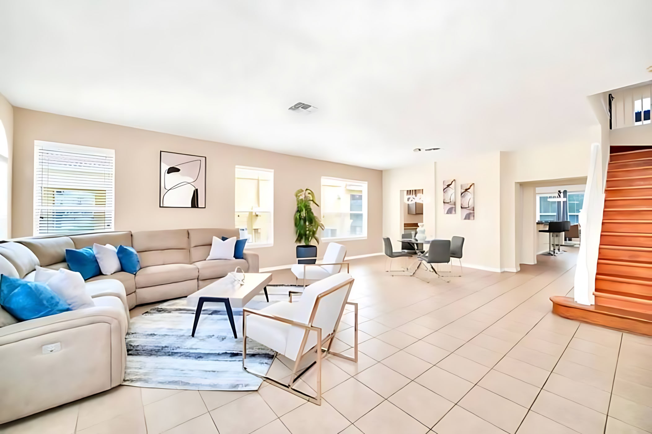 Expansive living area with light tiled floors, a large beige sectional sofa with blue and white pillows, a patterned rug, a glass-top coffee table, and white armchairs, with a view towards a dining area and staircase.