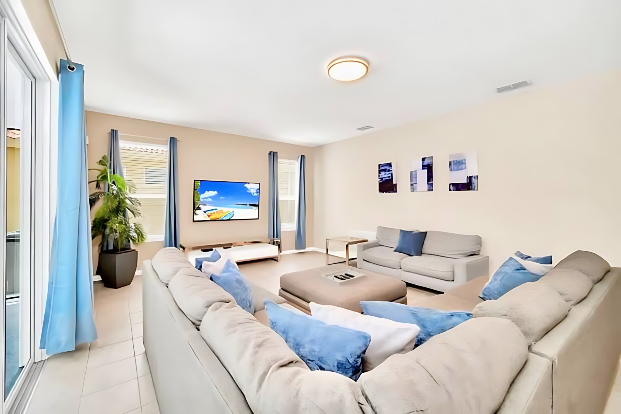 Modern living room featuring light tiled floors, a large L-shaped sectional sofa with blue and white pillows, a separate gray sofa, a wall-mounted TV, and blue curtains on windows and a sliding glass door.