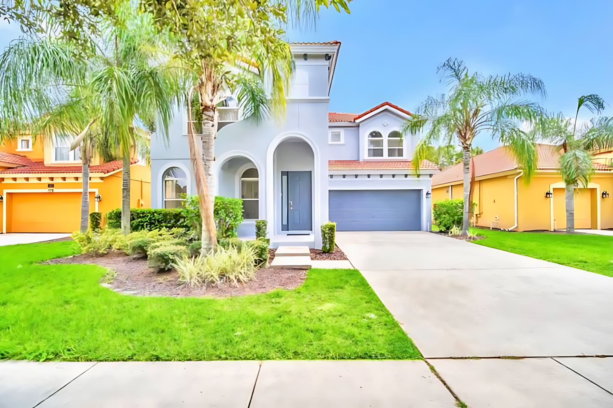 The front exterior of a modern two-story house features a light gray facade with arched entryways and a red tile roof. A well-maintained green lawn, palm trees, and a concrete driveway leading to a two-car garage complete the inviting curb appeal.