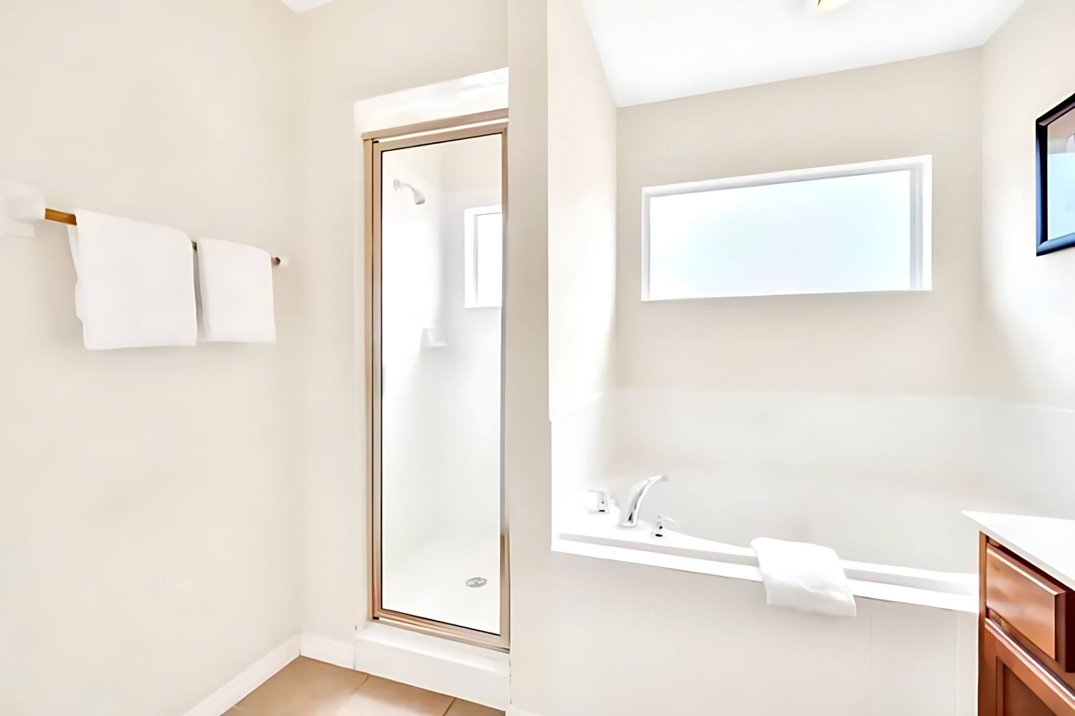 This bathroom view shows a built-in bathtub with a small window above, adjacent to a glass-enclosed walk-in shower, with white towels hanging on a rack.