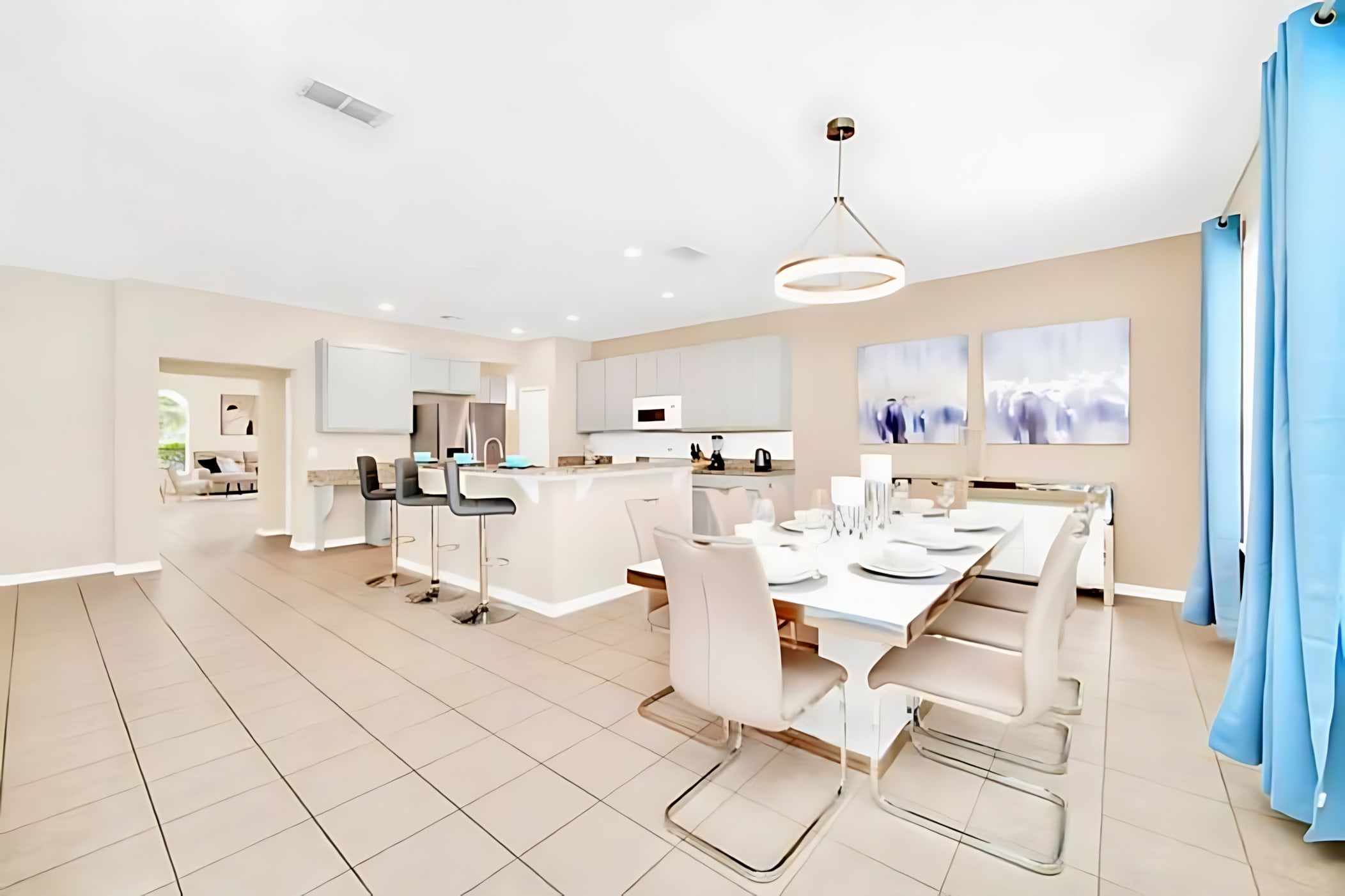 Open-concept dining area with light tiled floors, a rectangular white dining table with modern cream chairs, a contemporary chandelier, and a view into the adjacent kitchen with light gray cabinets.