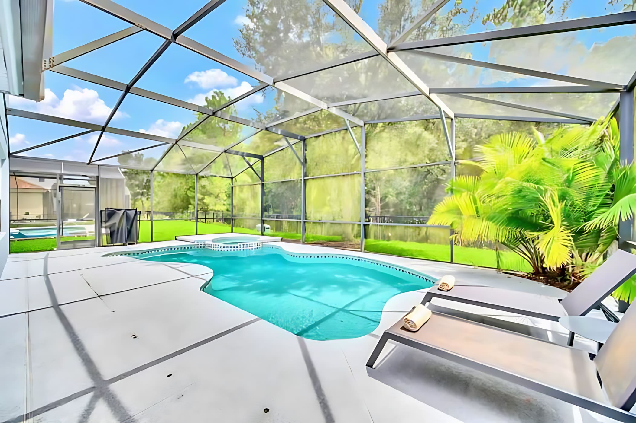A wide shot of the screened-in outdoor pool area showcases a freeform pool and hot tub, with two modern lounge chairs on the white concrete deck. The surrounding lush greenery and clear blue sky are visible through the enclosure.