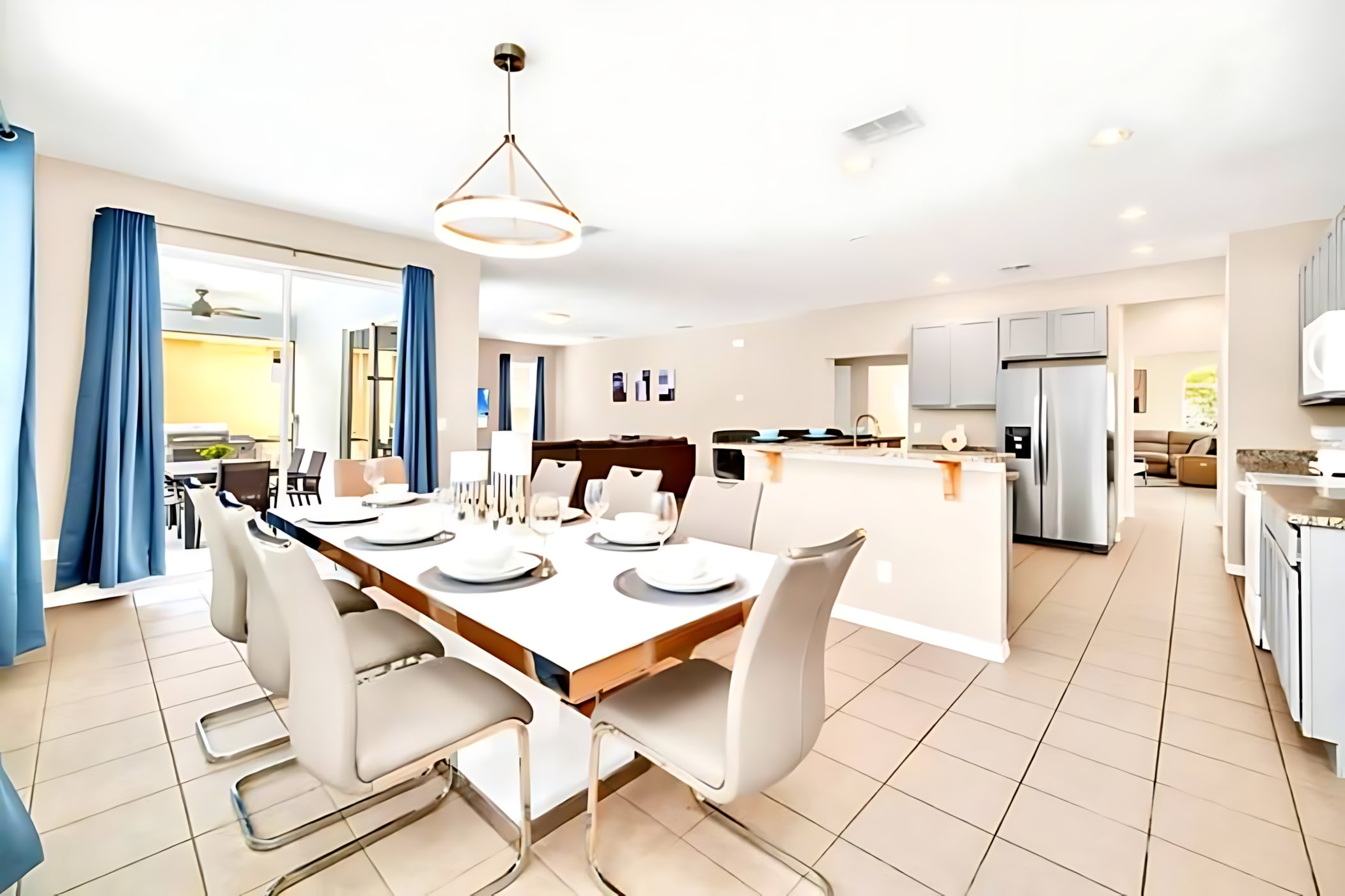 An open-concept dining area features a modern white rectangular dining table with six gray chairs, illuminated by a contemporary chandelier. The adjacent kitchen with stainless steel appliances and a breakfast bar is visible in the background.