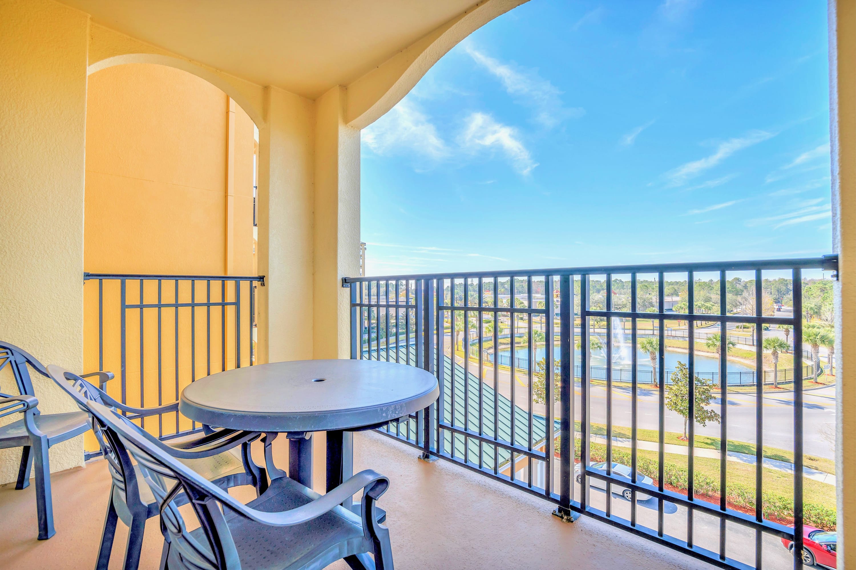 A private balcony offers outdoor seating with a small round table and two chairs, overlooking a resort landscape with a fountain and other buildings under a clear blue sky.