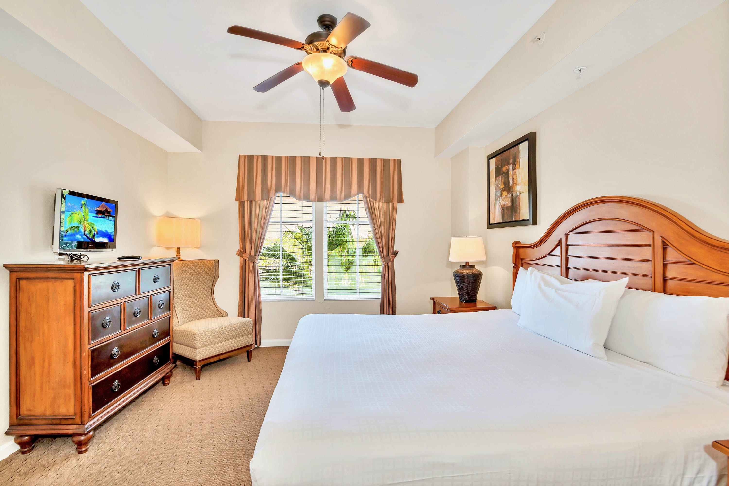 Spacious master bedroom with a king-sized bed, dark wood headboard, ceiling fan, and a large window with striped valance and views of palm trees. A wooden dresser with a TV and an armchair are also visible.