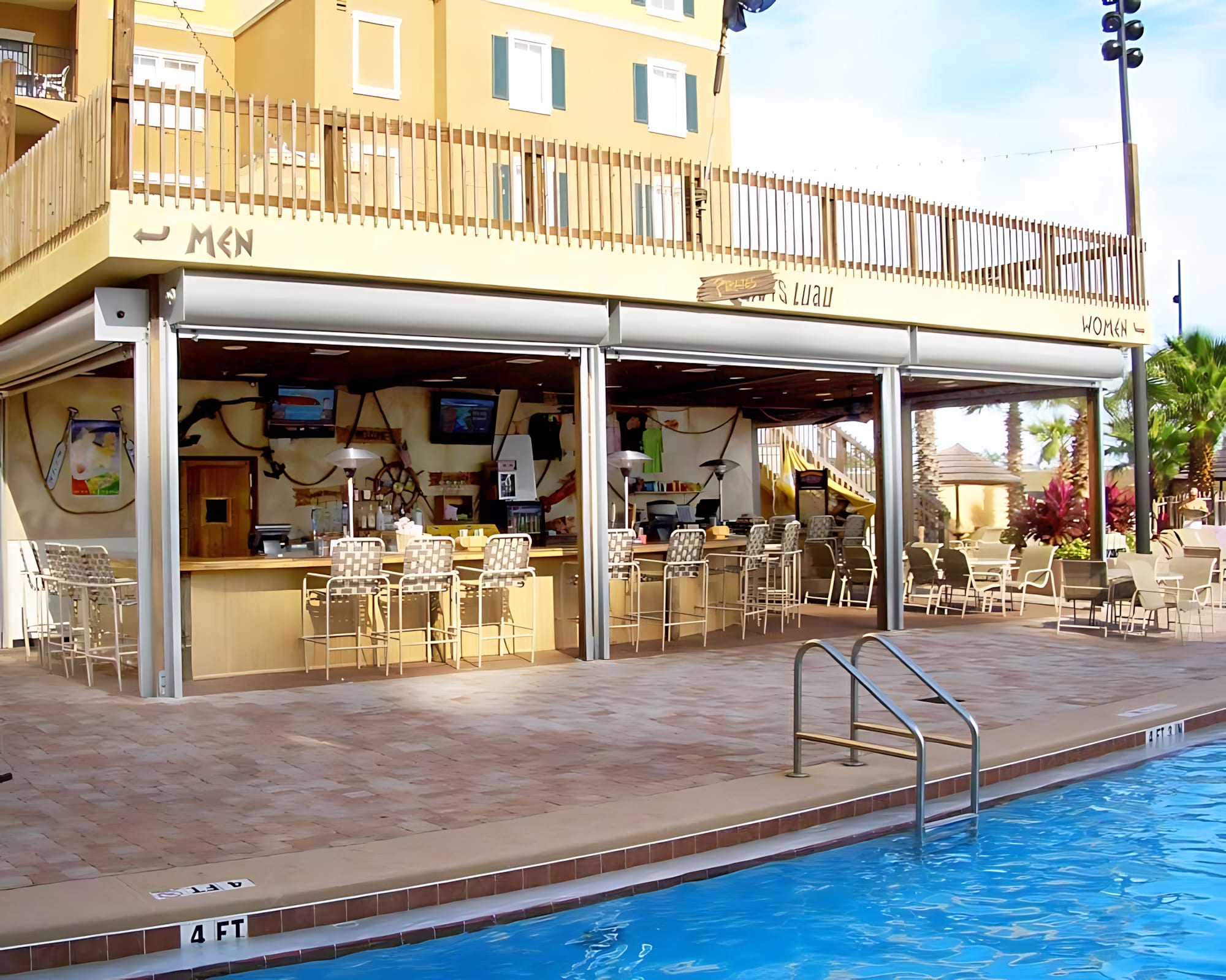 An outdoor pool bar with a light-colored wooden counter, bar stools, and multiple TVs, situated next to a swimming pool with a brick-patterned deck. The bar is open-air with roll-up doors and a second-story balcony above.