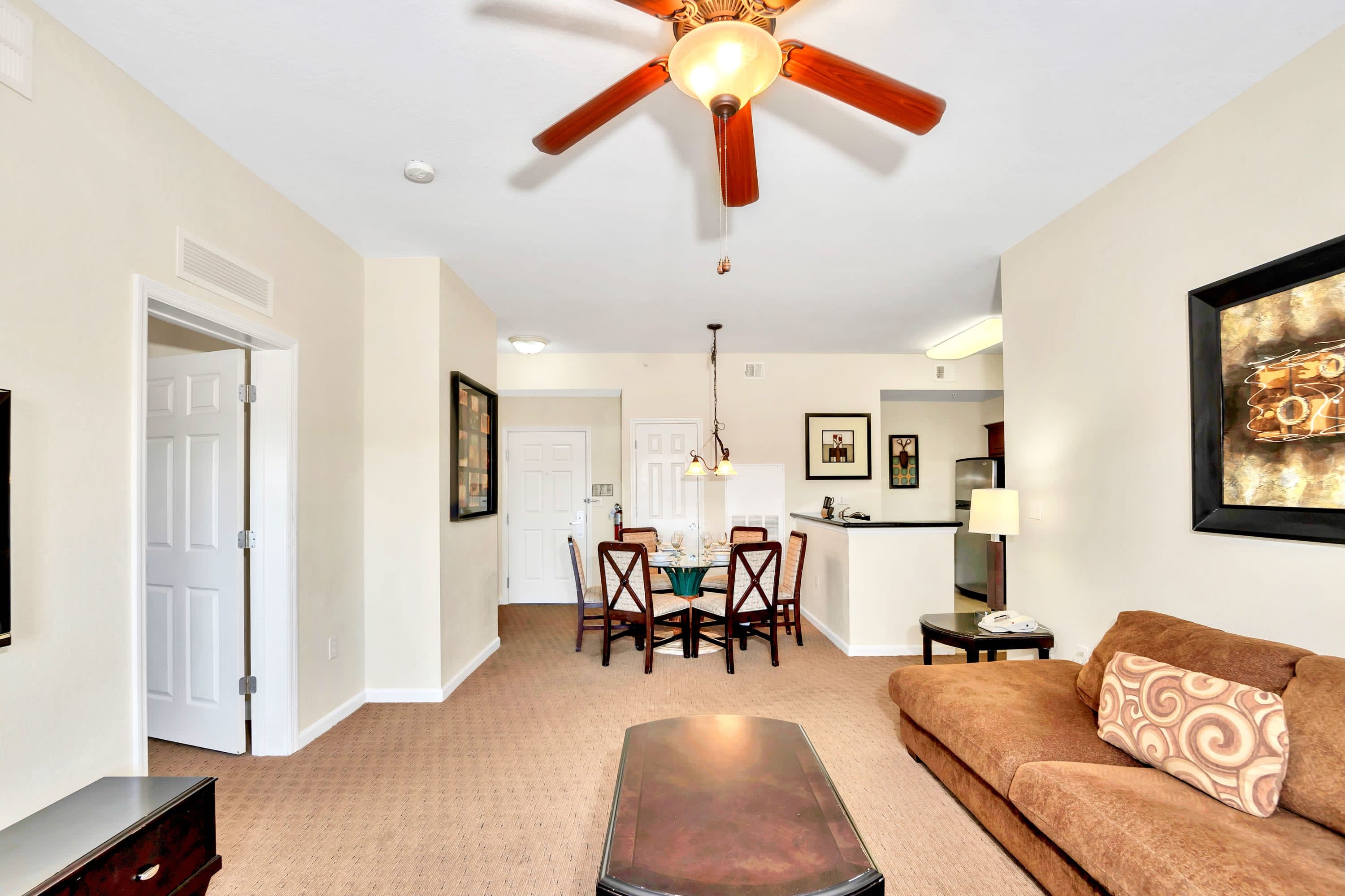 An open-concept living space shows a brown sectional sofa and coffee table in the foreground, leading to a dining area with a glass table and chairs, and a glimpse of the kitchen beyond.